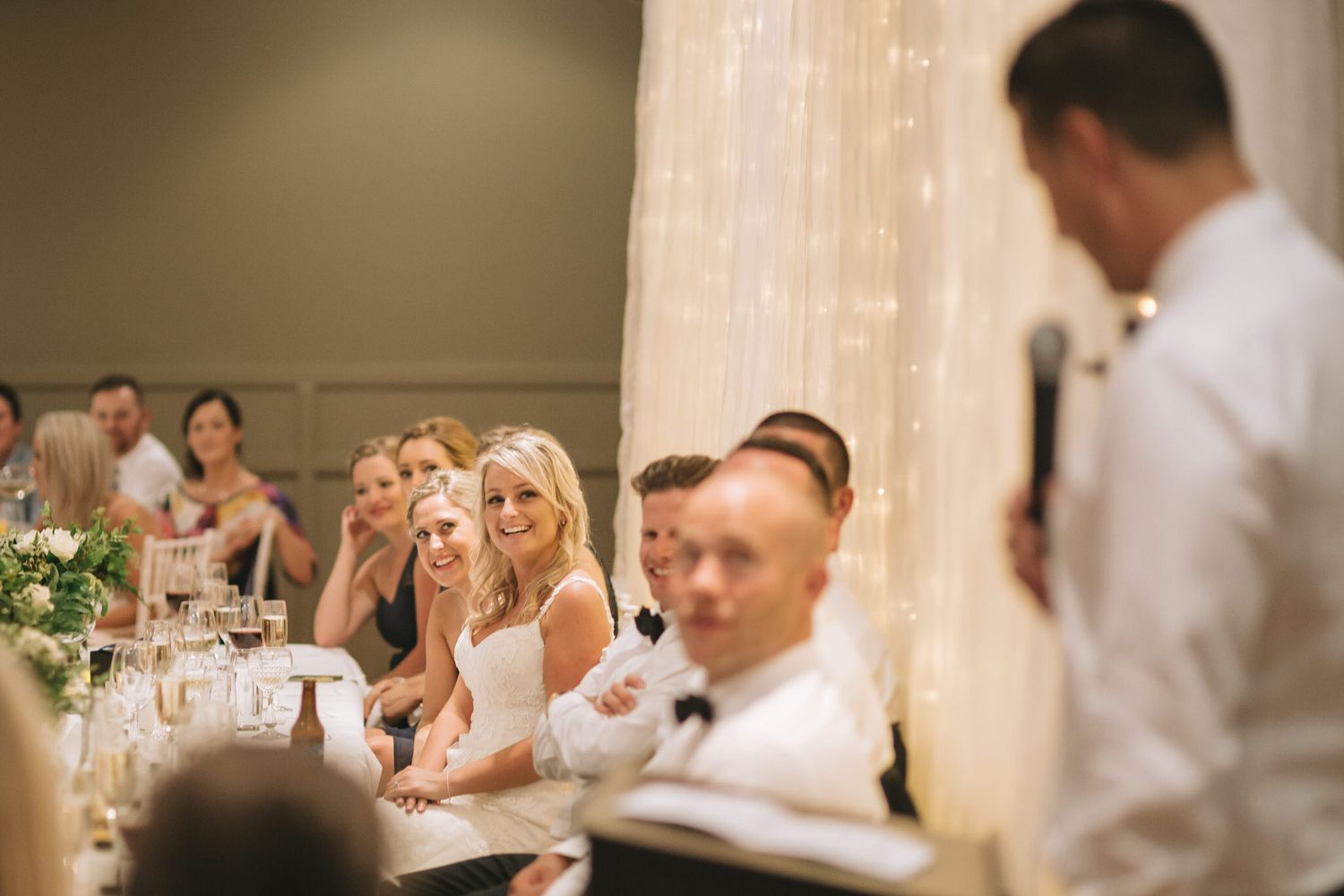 Wedding guests at a reception dinner table listen and smile during a speech at an indoor celebration.