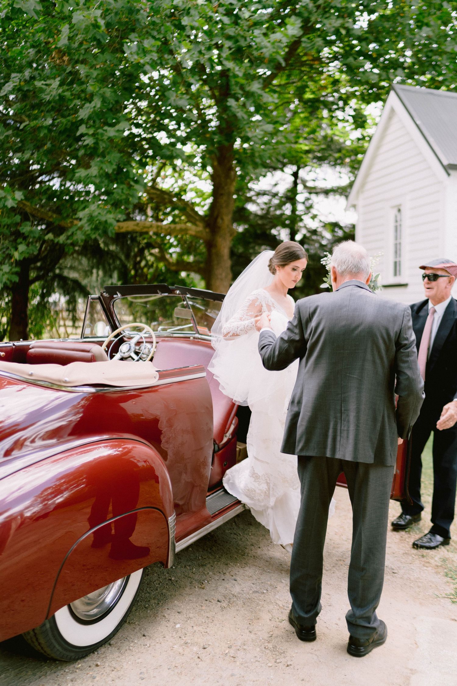 Wedding guests gather around a classic burgundy convertible car parked under green trees.
