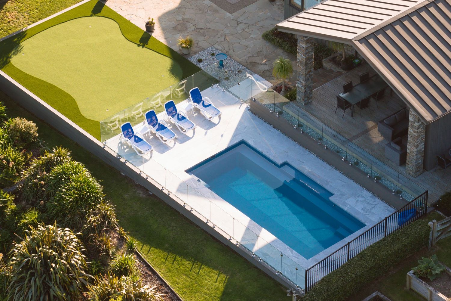 Aerial view of a luxury home with a rectangular pool, blue lounge chairs, and a putting green.