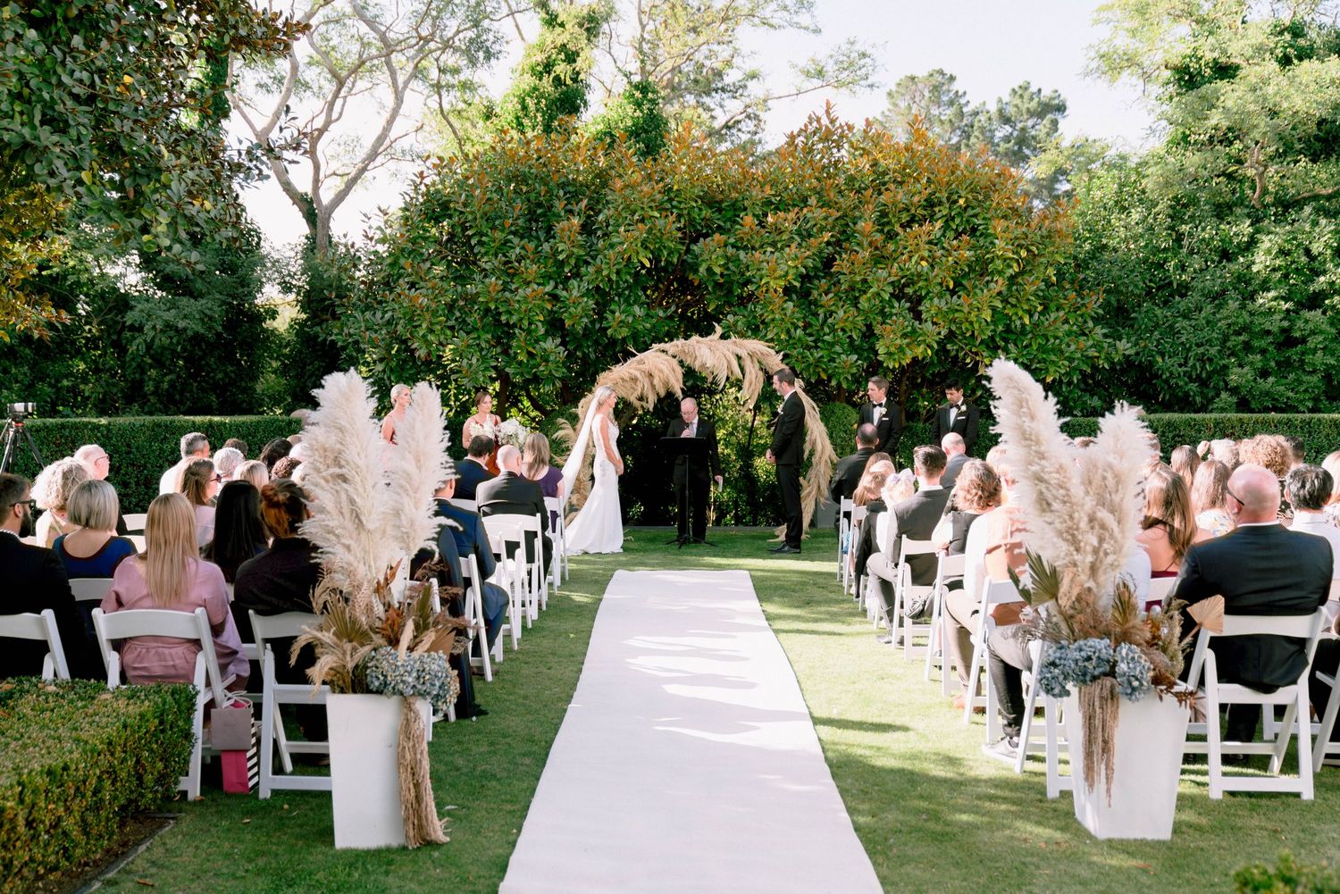 Outdoor wedding ceremony setup with a floral arch and white chairs lining the grass aisle with pampas grass decorations.