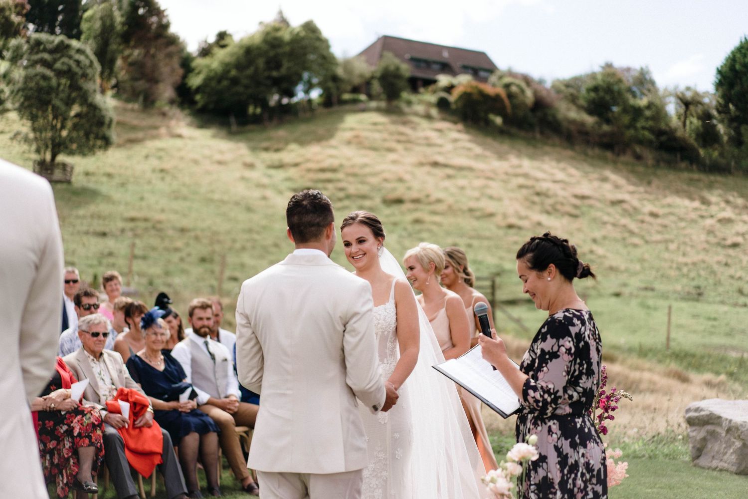 A wedding ceremony taking place outdoors on a hillside with guests seated on white chairs.
