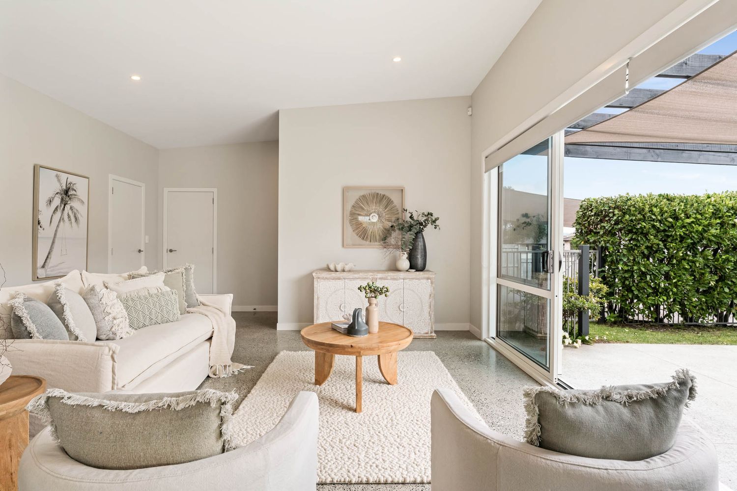 Modern coastal living room with white sofa, gray chairs and sliding glass doors opening to outdoor patio with greenery.
