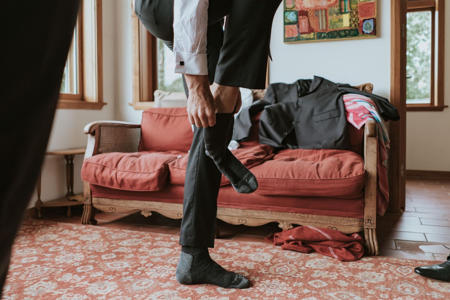 Someone in black pants and socks stands near a pink couch with scattered clothes in a sunlit room.