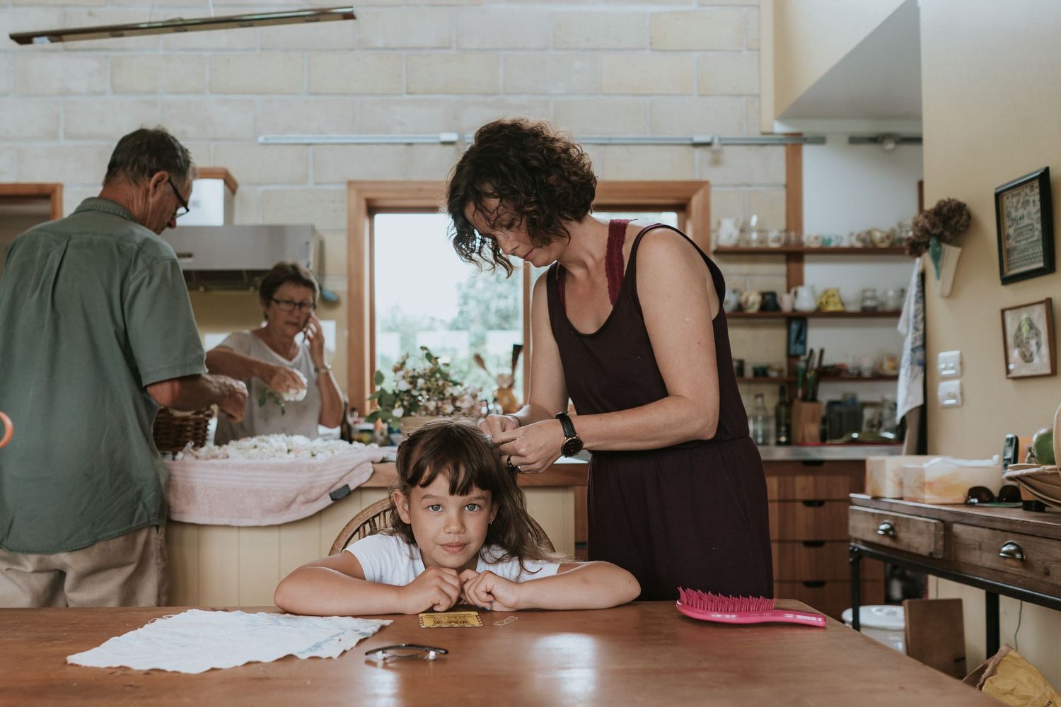 A family bakes together in a cozy kitchen with wooden shelves and natural light streaming through the window.