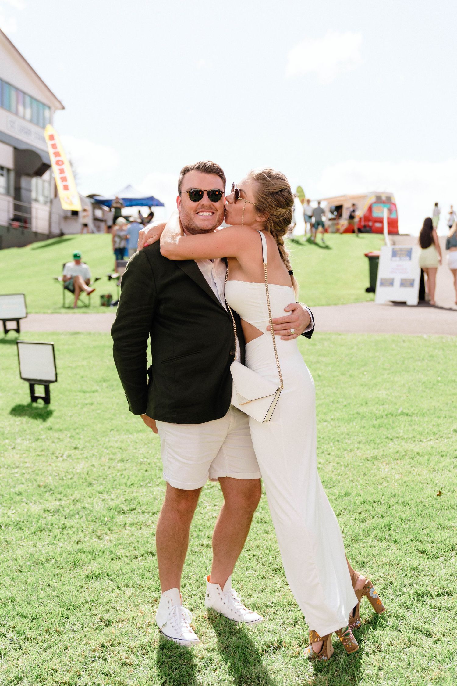 A couple embraces on a sunny lawn during an outdoor event with tents and guests in the background.