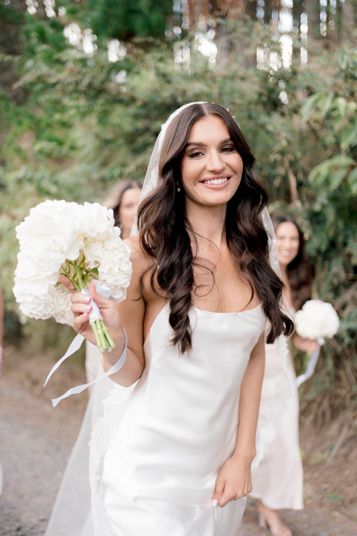 A bride in a sleeveless white dress holding a white bouquet against a forest backdrop.