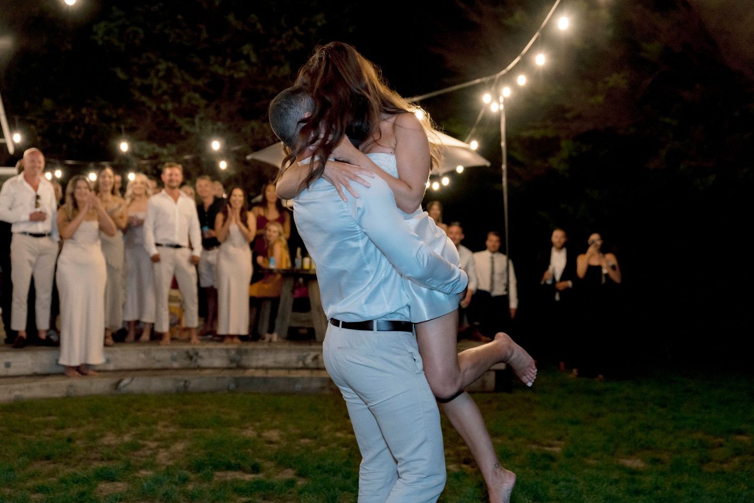 Couple dancing under string lights at outdoor evening wedding reception.
