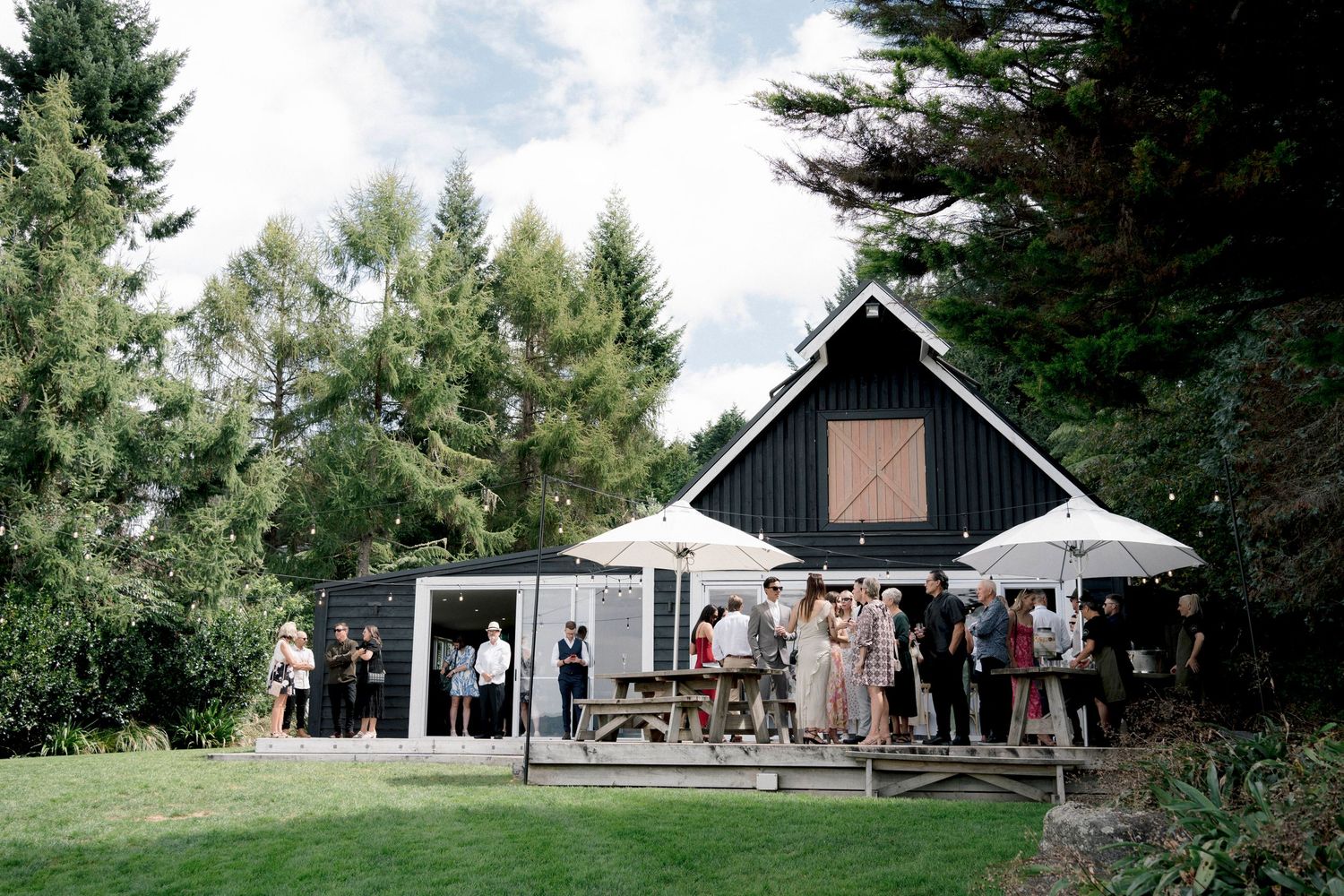 A modern black and white barn-style venue with guests gathered on the patio surrounded by evergreen trees.