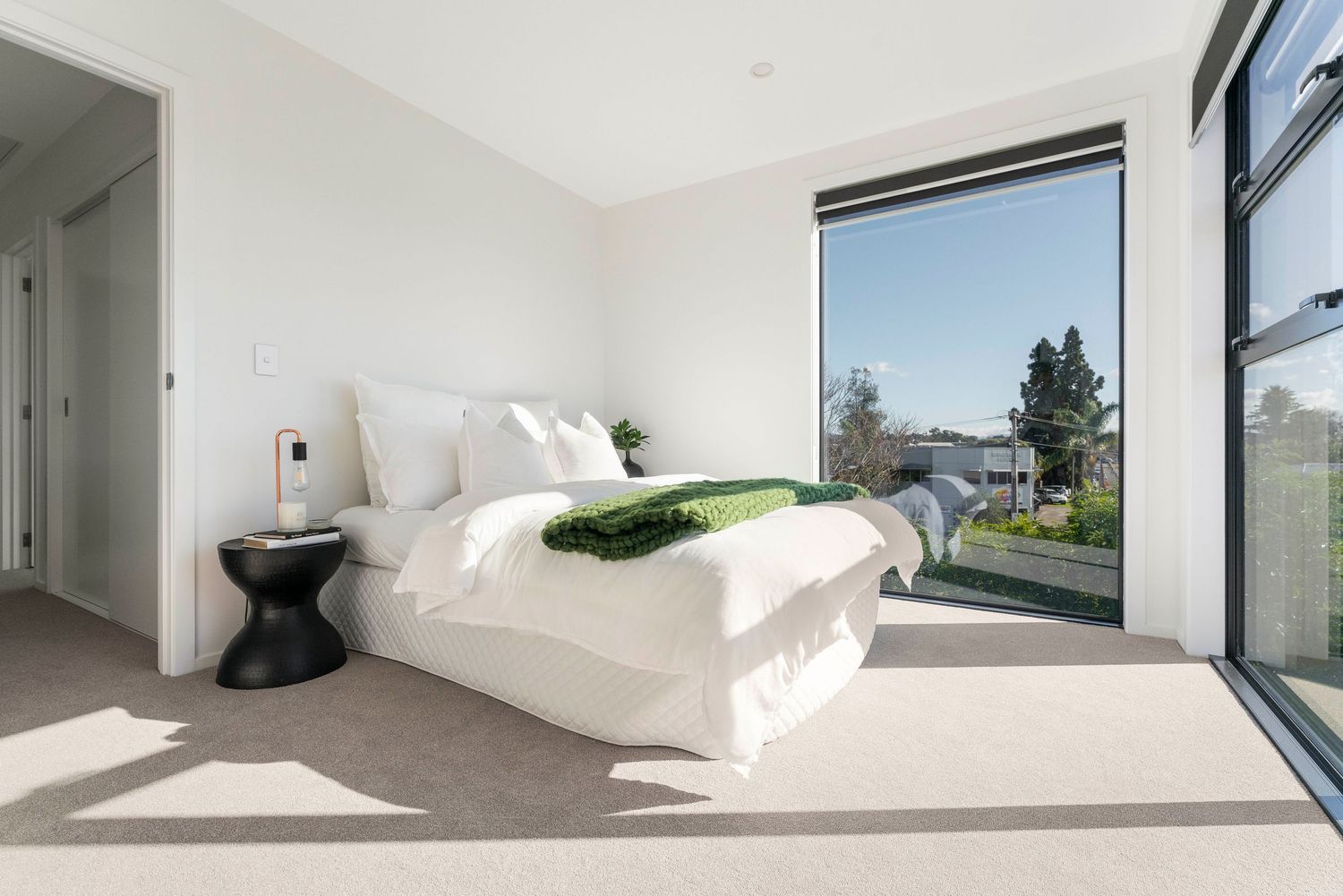 Modern bedroom with white bedding features large windows, natural light and minimalist decor in a contemporary space.