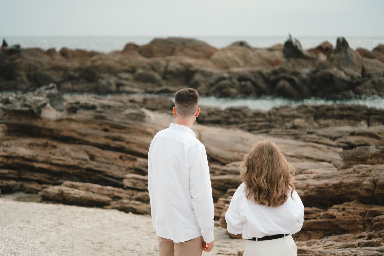 Two people sit together on rugged coastal rocks as waves crash against the rocky shoreline at sunset.