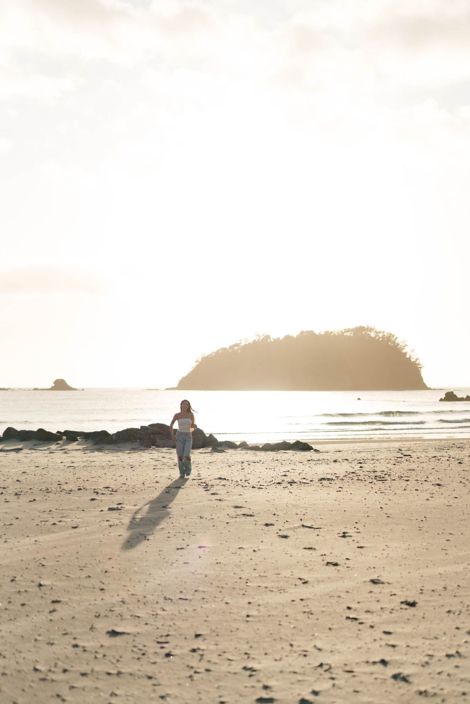 A solitary figure walks along a misty beach at sunset casting a long shadow on the sand.