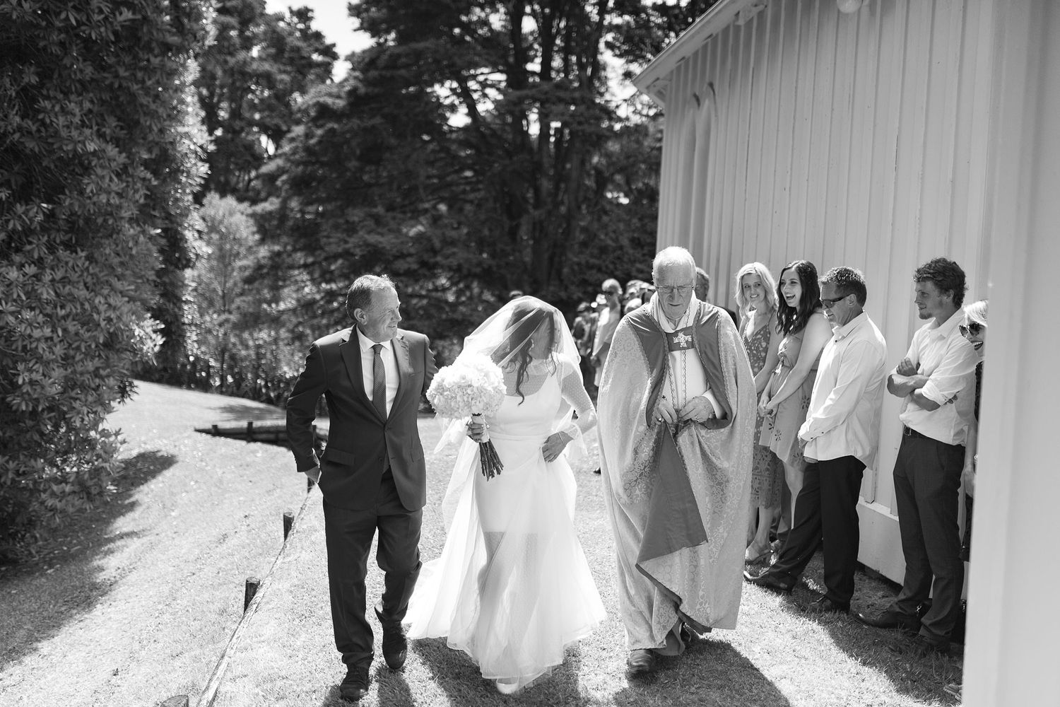 A wedding party walks along a path next to a white building in this black and white photograph.