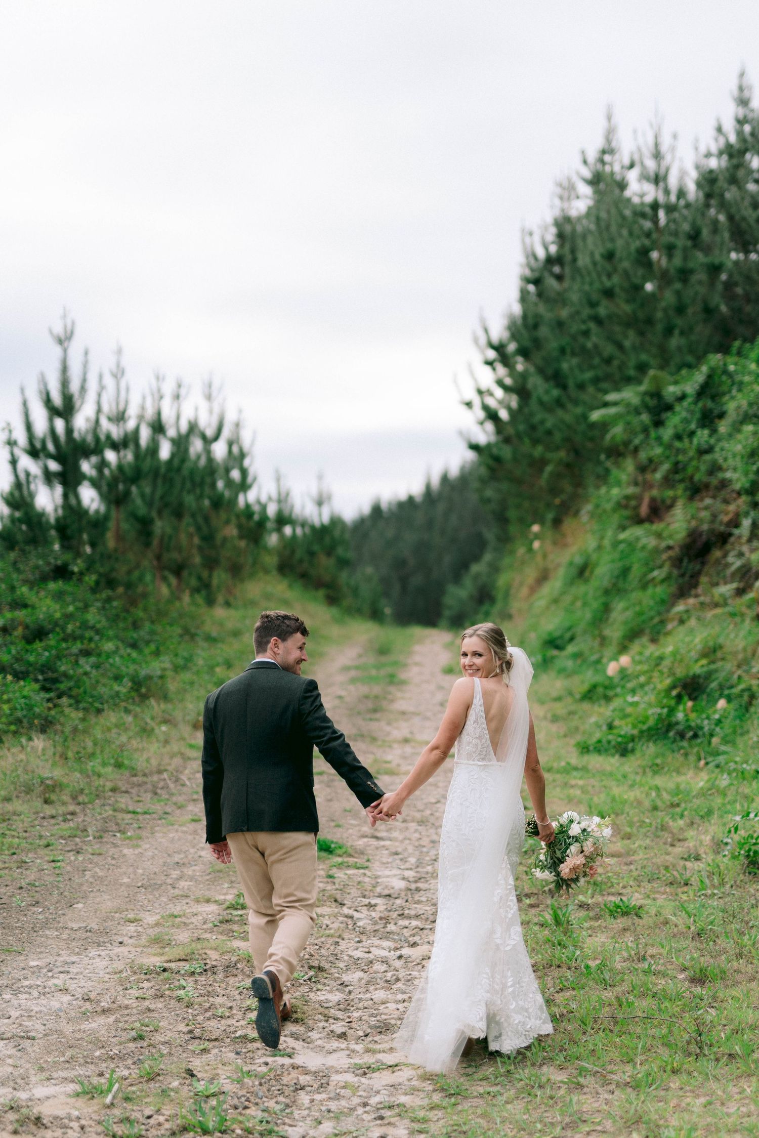 A couple walks hand in hand down a forest path with evergreen trees on a cloudy day.