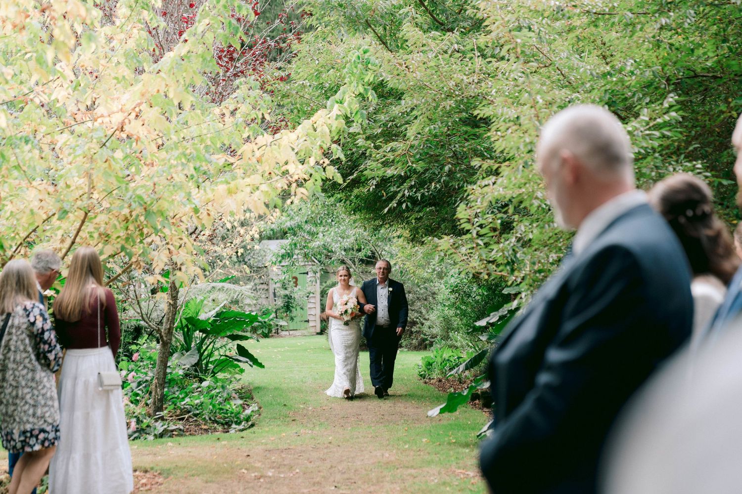 Outdoor garden wedding ceremony with guests watching couple exchange vows among lush greenery.