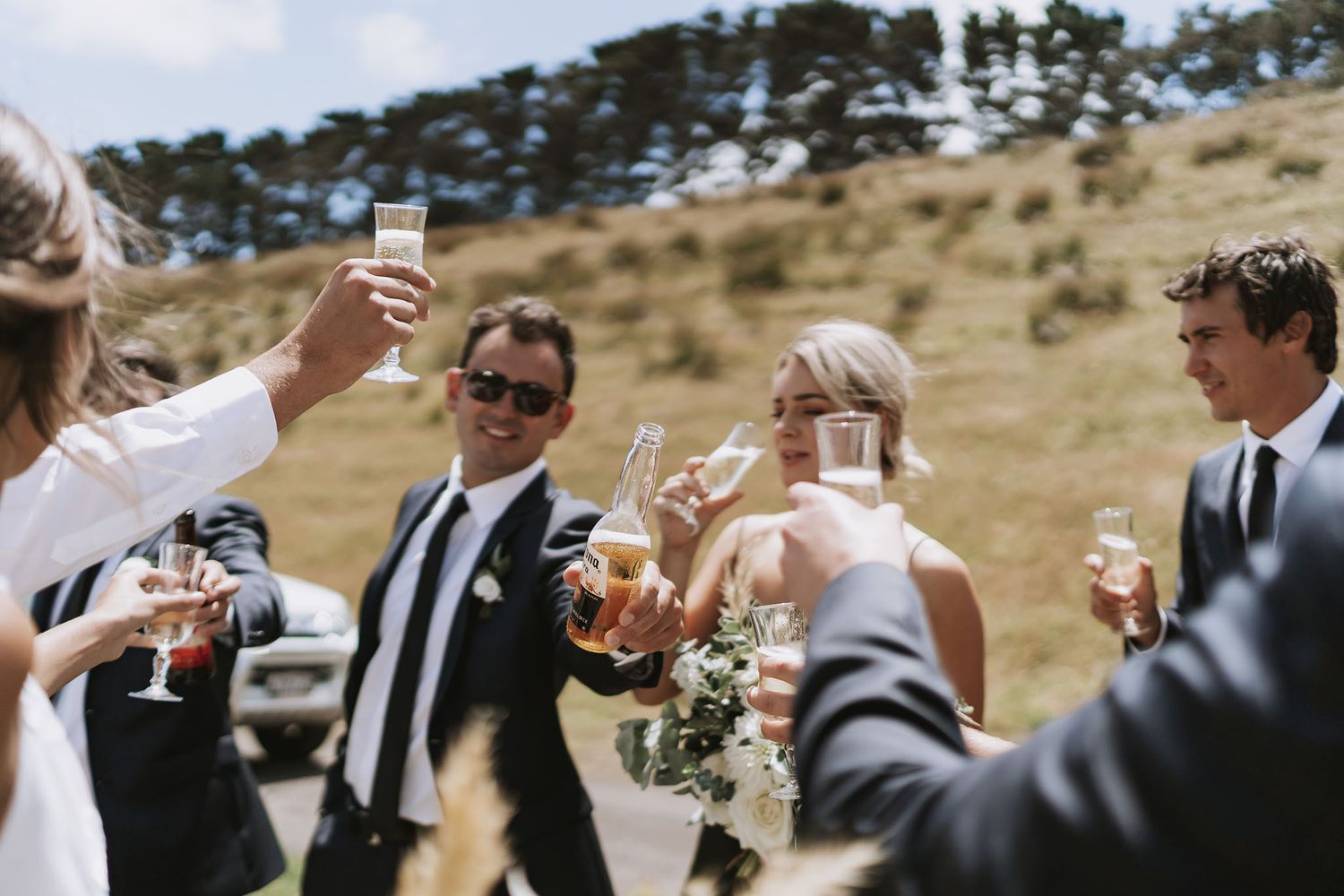 Wedding guests dressed in formal attire raise champagne glasses for a toast on a sunny hillside during an outdoor celebration.