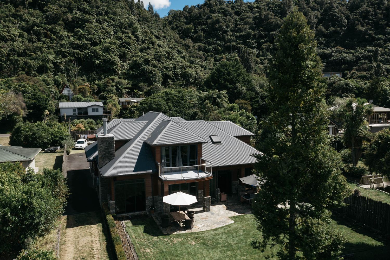 Modern luxury house with dark metal roof and outdoor patio surrounded by lush green forest landscape viewed from above.