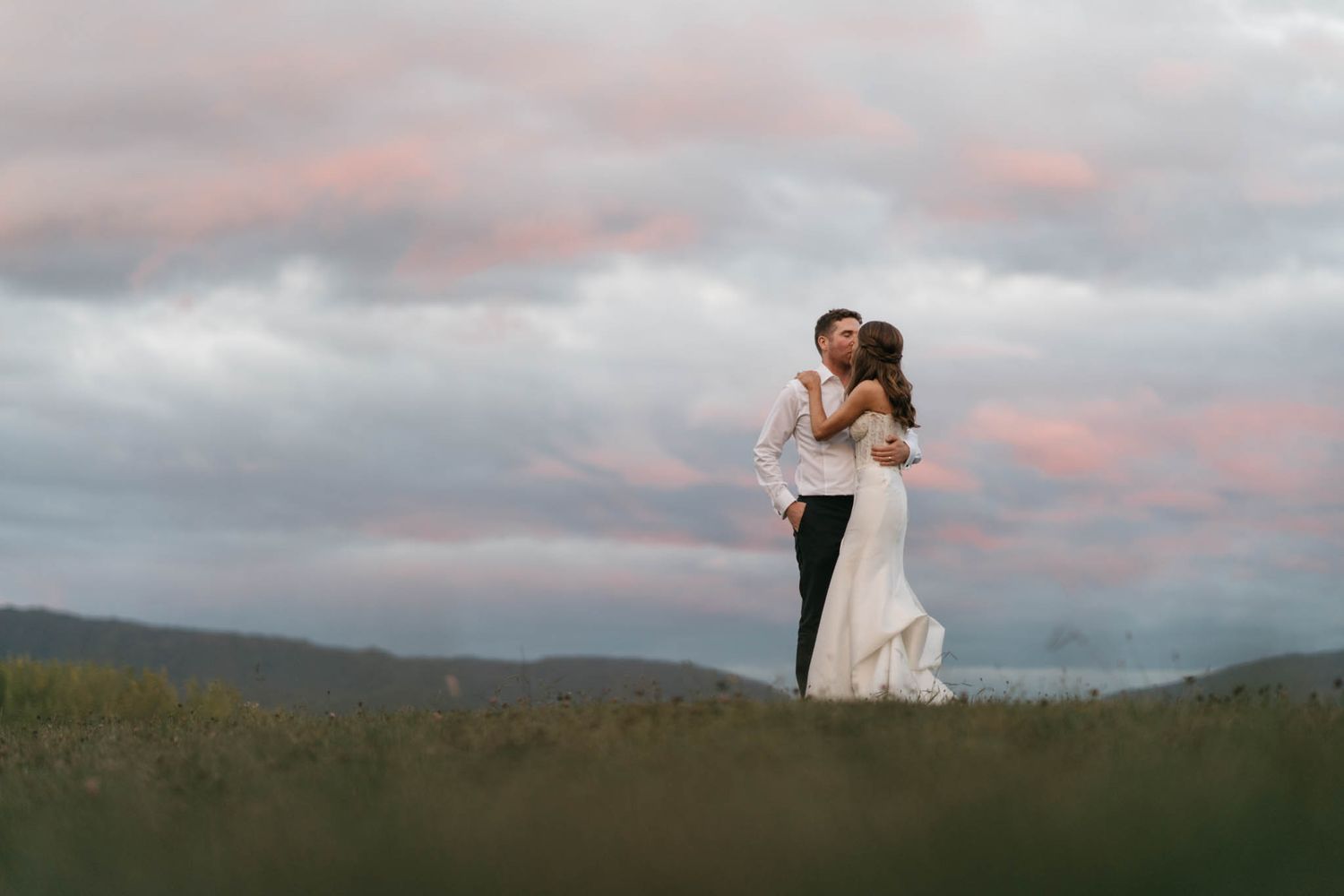 A couple embraces on a hilltop against a dramatic cloudy sunset sky with mountains in the background.