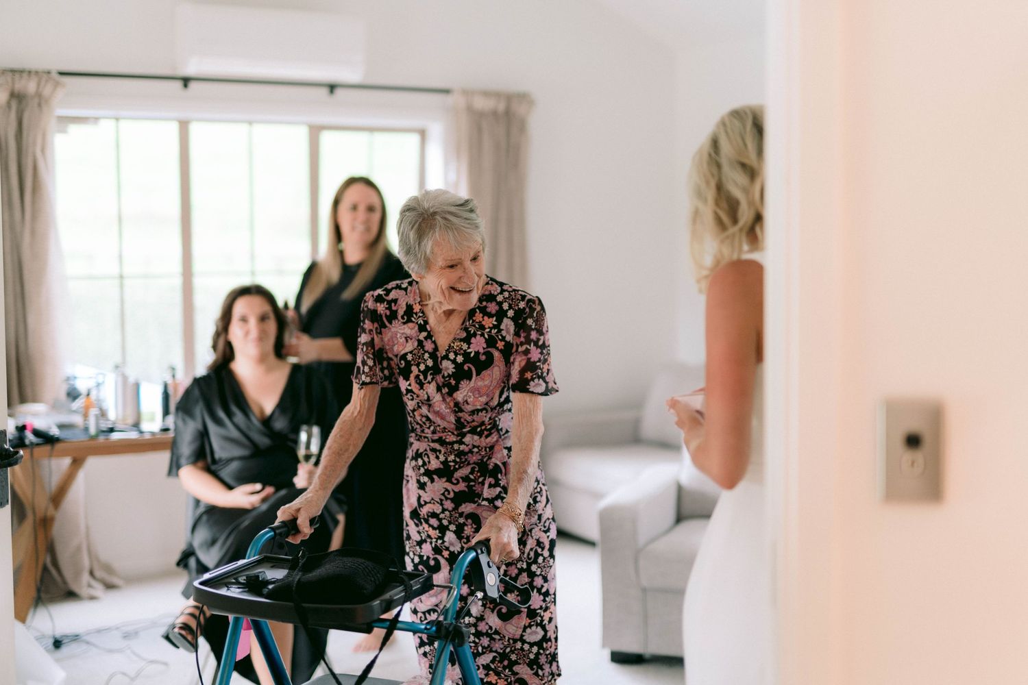 People gather in a bright interior room with white walls and natural lighting during wedding preparations.