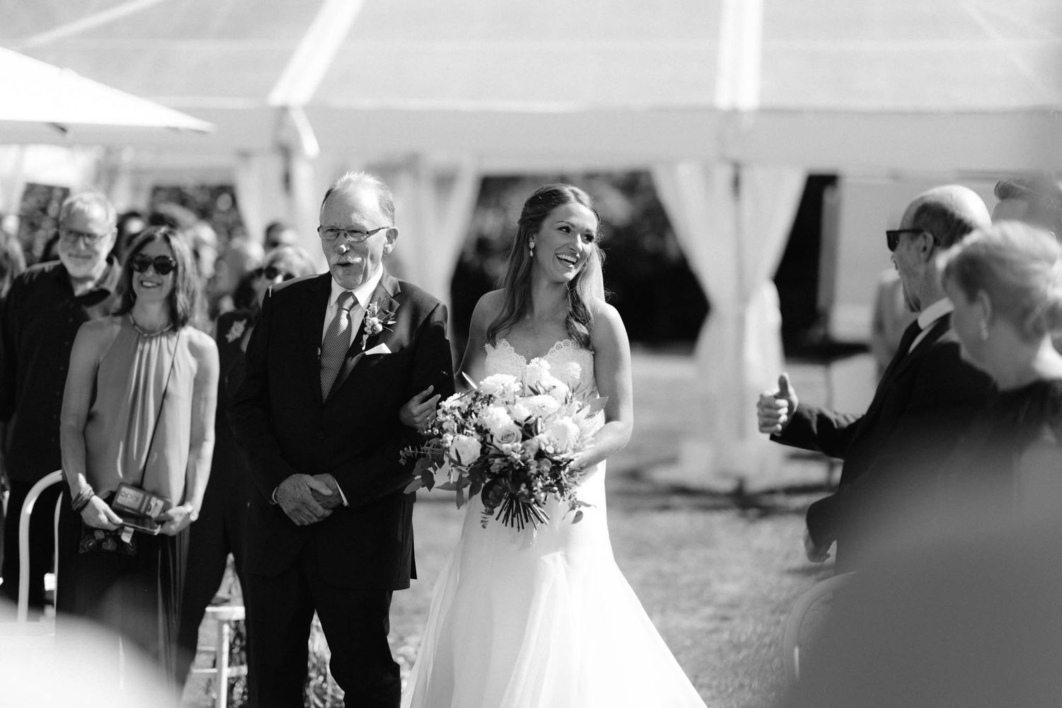 Black and white photo of a traditional outdoor wedding ceremony under a tent with flowers and gathered guests.