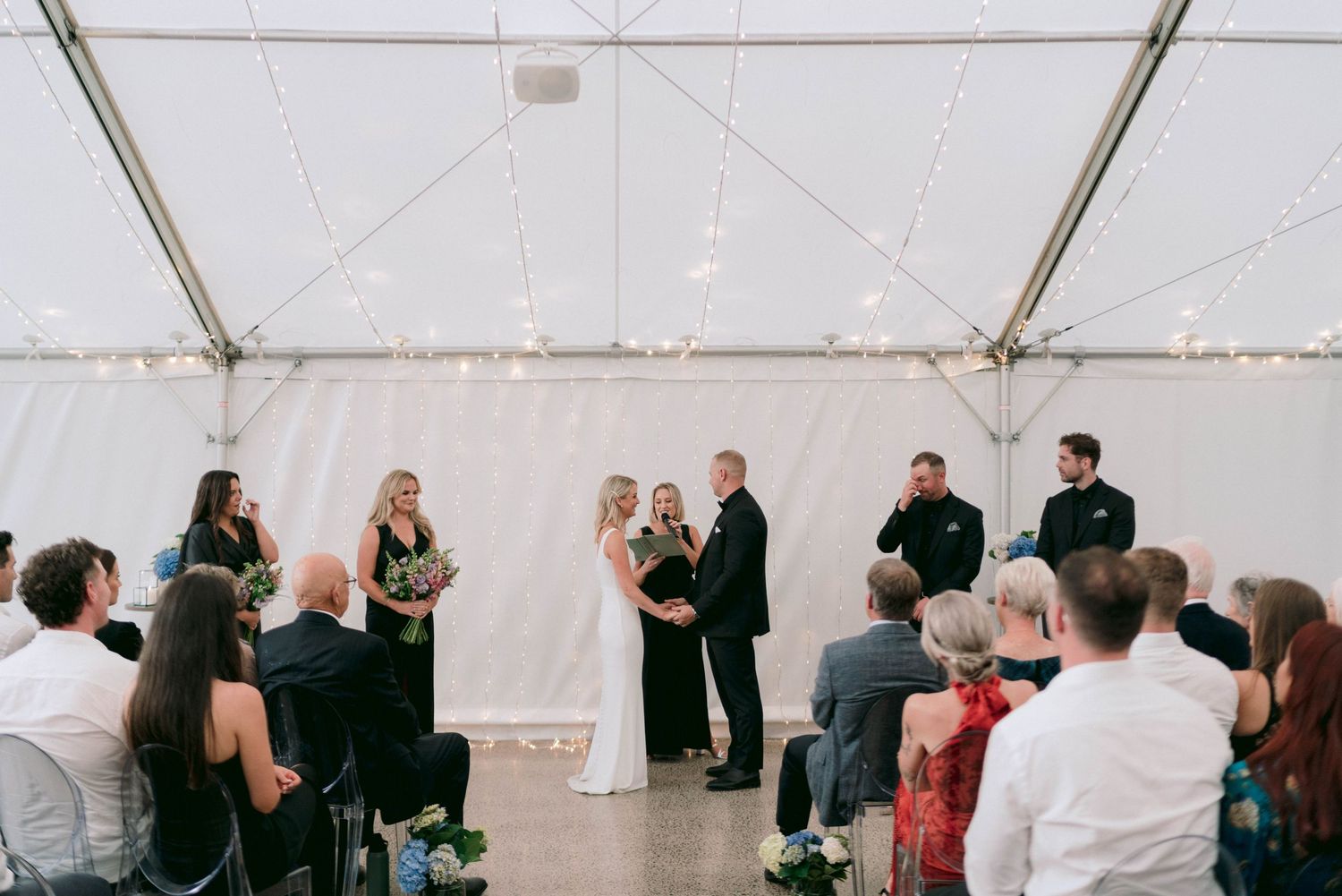 Wedding guests mingle and celebrate inside a white tent decorated with overhead string lights.