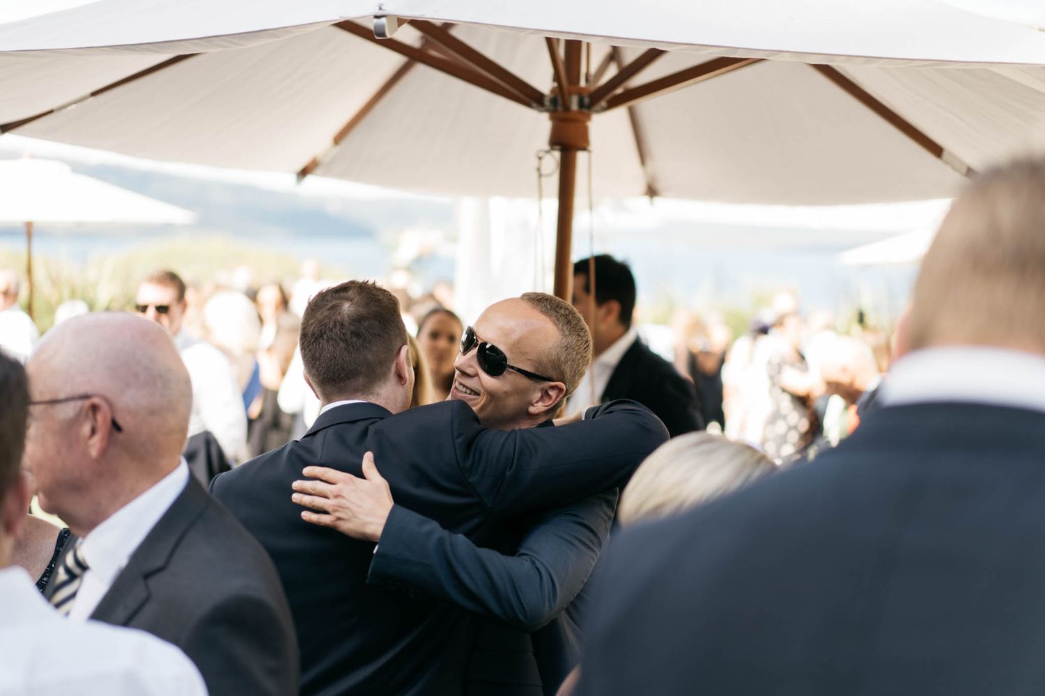 A group of formally dressed guests gather under a white outdoor umbrella during a daytime event.