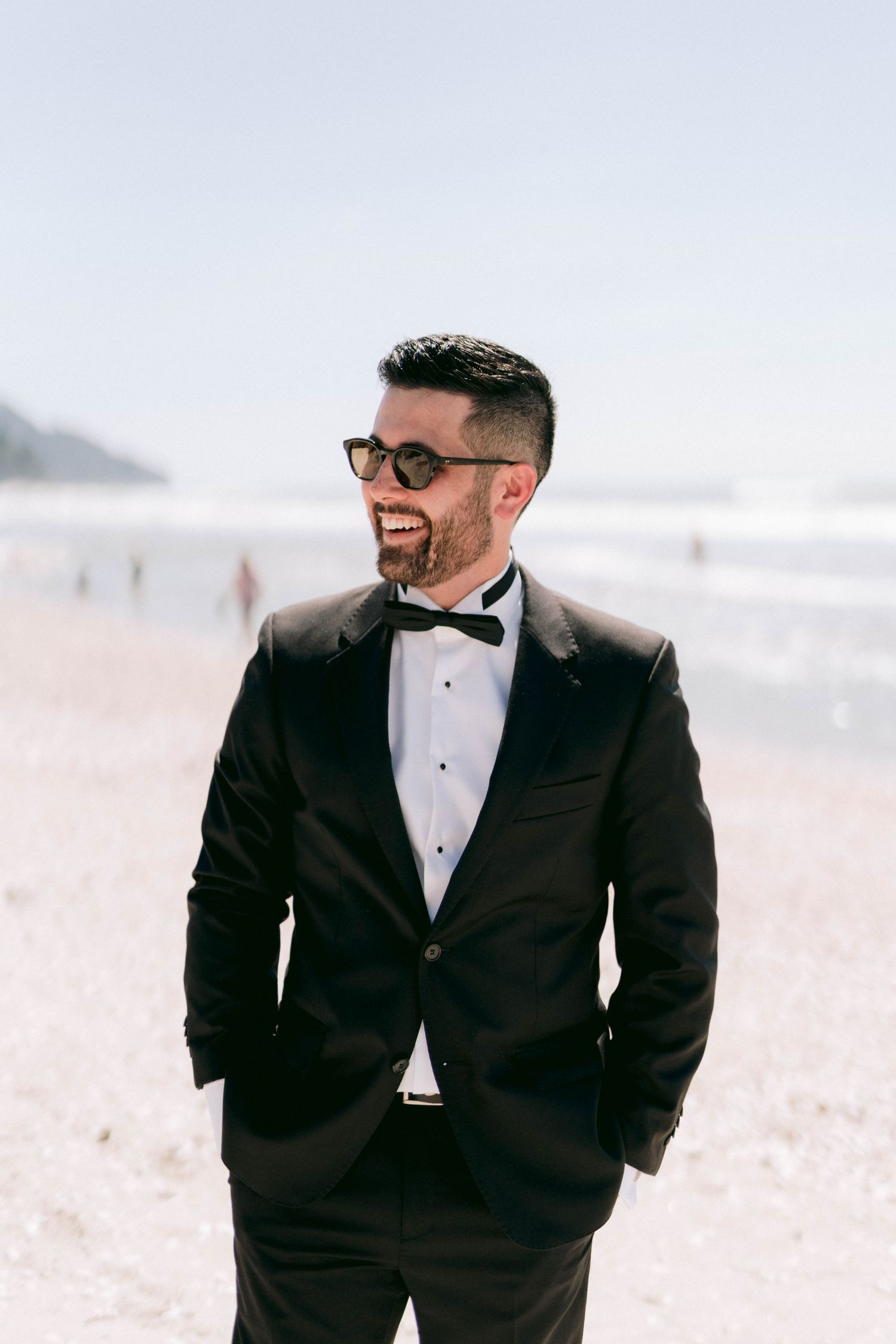 Portrait series of a groom in glasses and tuxedo laughing joyfully during beach wedding photoshoot.