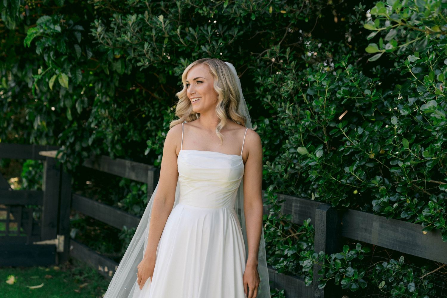 Woman in flowing white summer dress poses in a lush garden setting with rustic wooden fence.