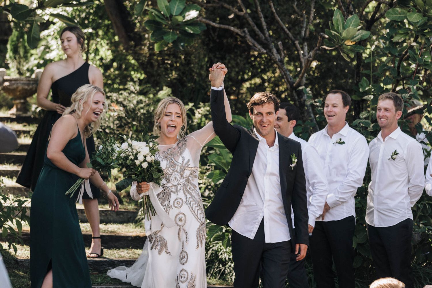 Wedding party celebrates on garden steps while guests throw confetti during joyful outdoor celebration.