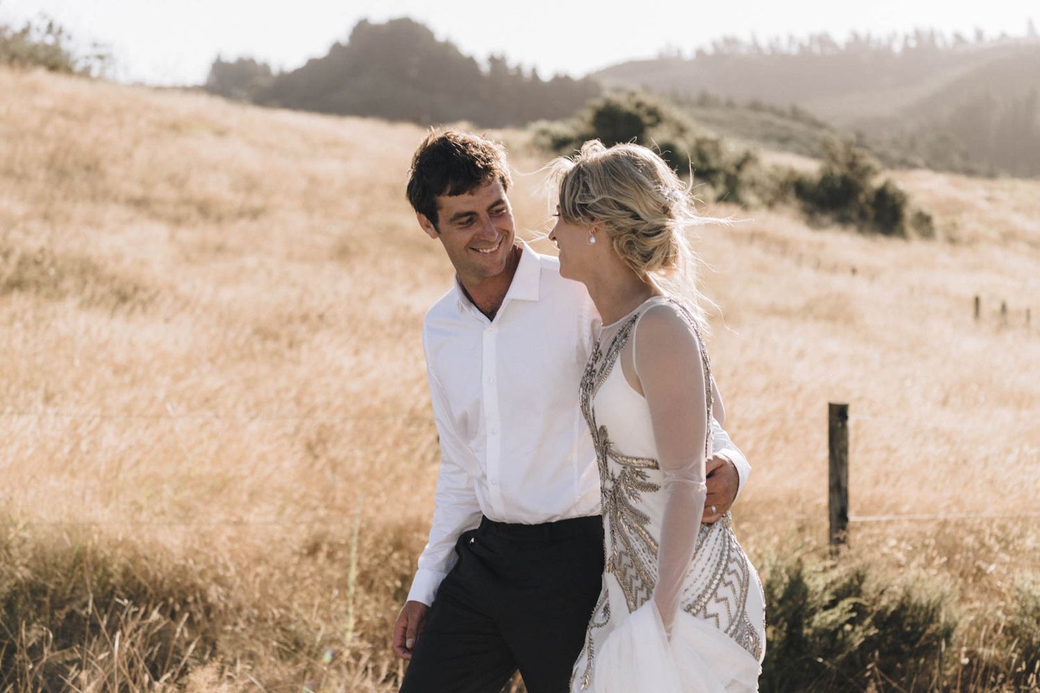 A couple in wedding attire share an intimate moment in a golden sun-lit field with rolling hills in the background.
