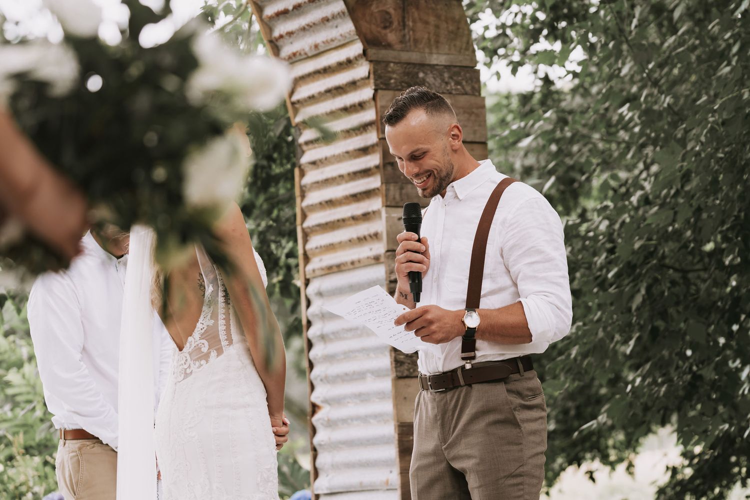 A white-clothed couple stands intimately together outdoors during a rustic wedding celebration.