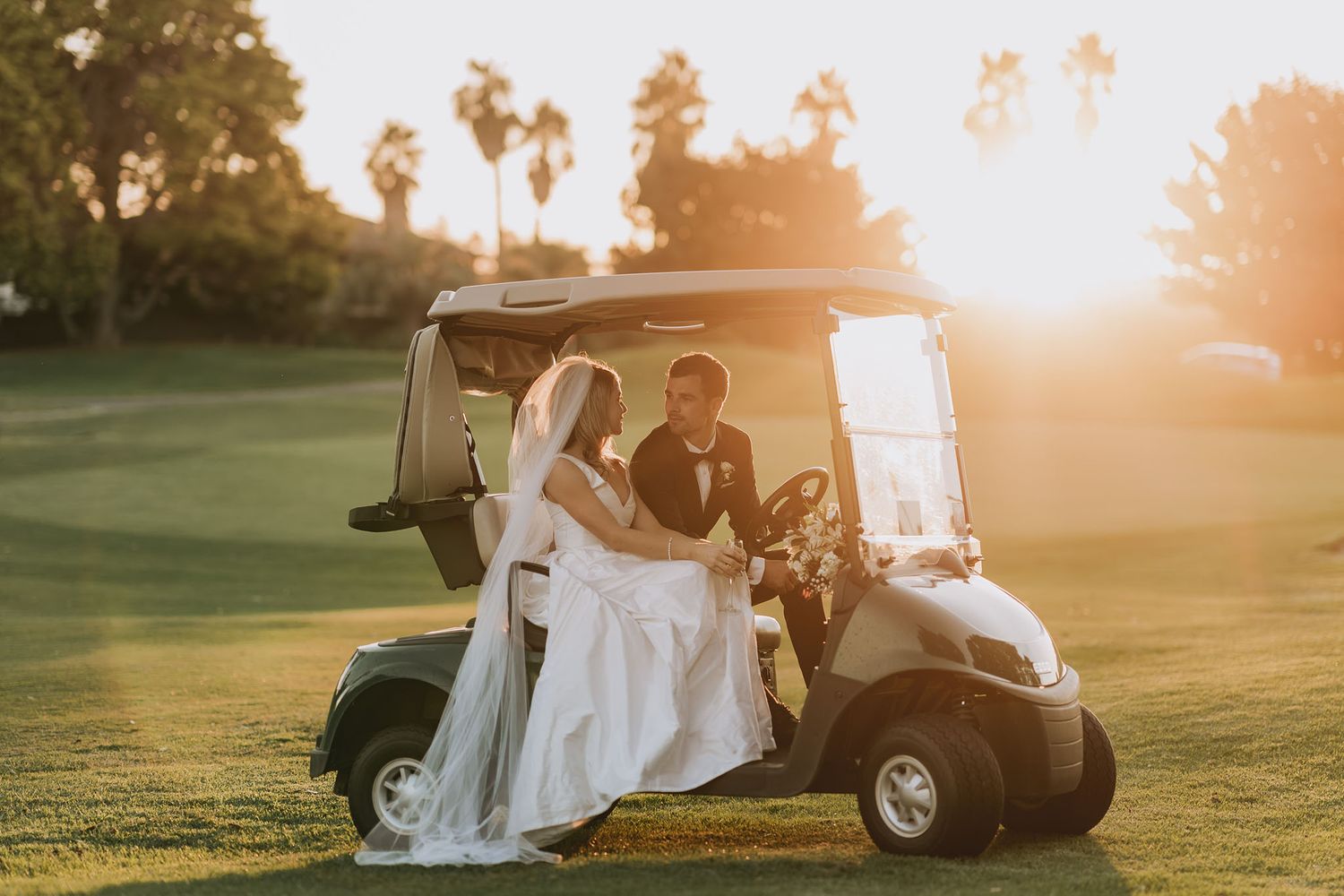 Couple in wedding attire sitting in golf cart on green course during golden hour sunset.