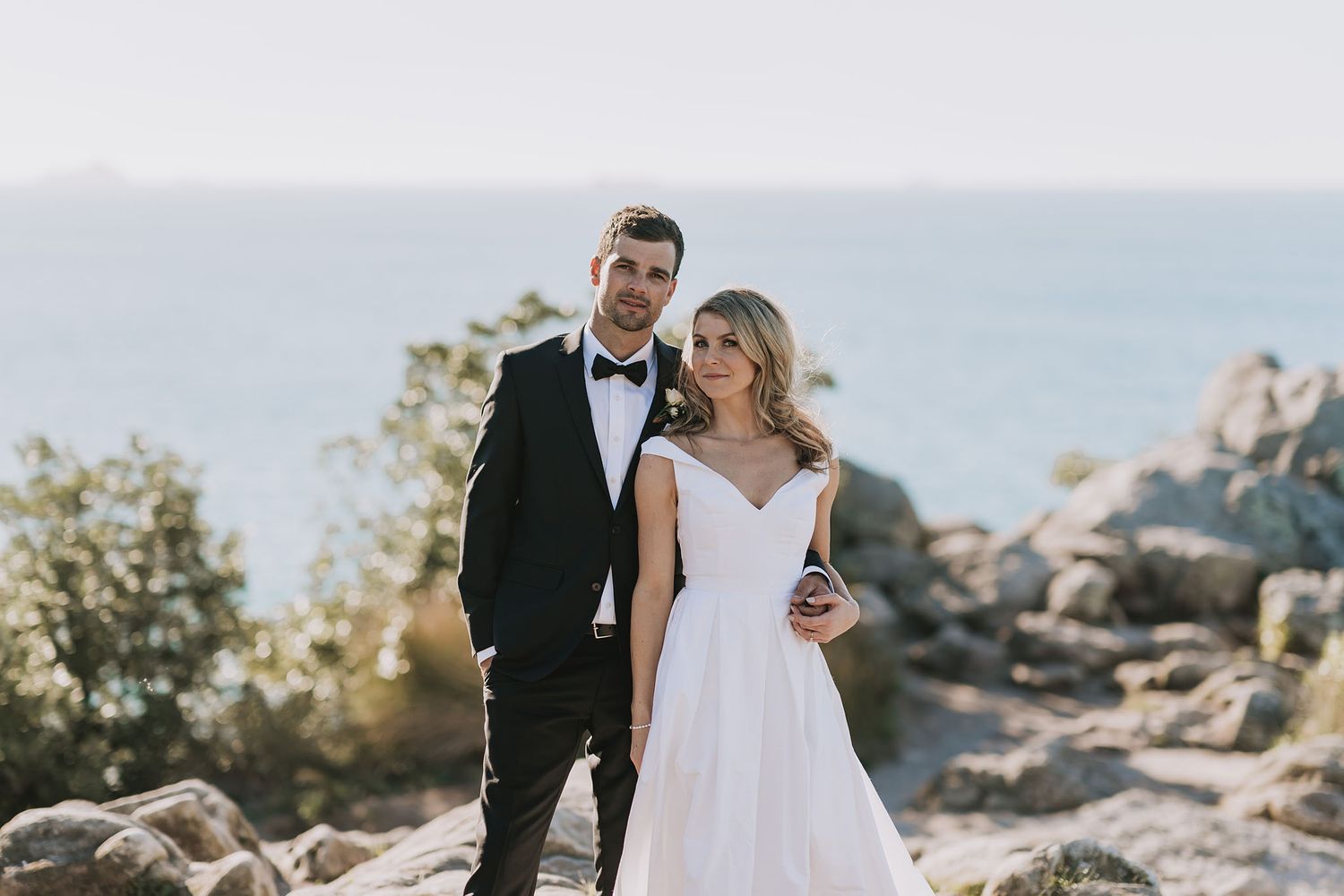 A couple in wedding attire stands on a rocky cliff overlooking mountains and wilderness at sunset.