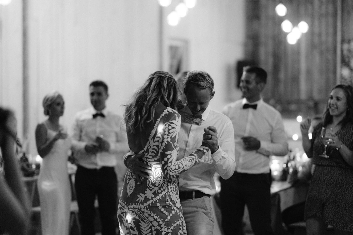 Black and white photo of wedding reception guests dancing and celebrating inside an elegant venue with blurred lights.