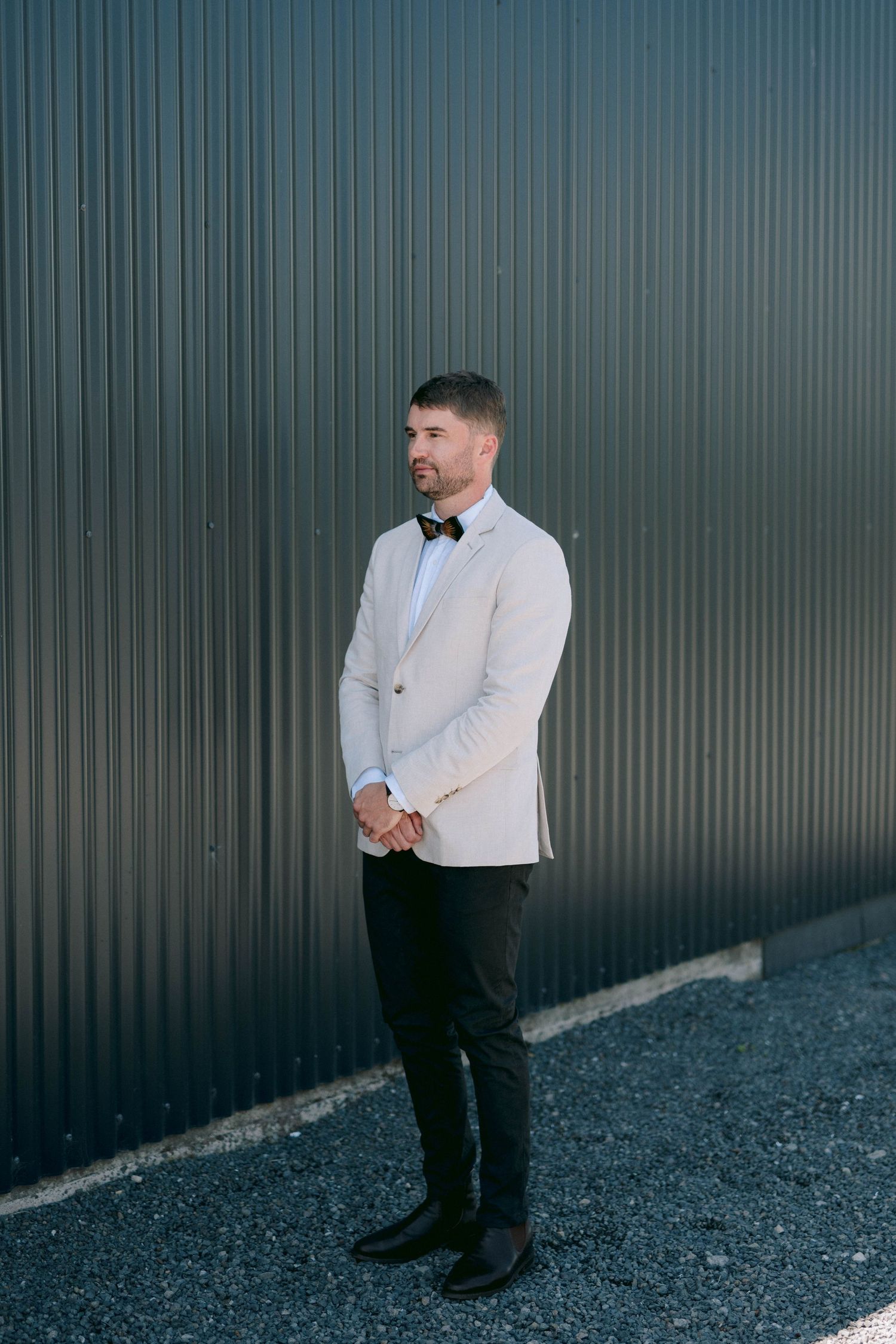 Groom wearing light colored jacket and bow tie stands against industrial metal wall background.