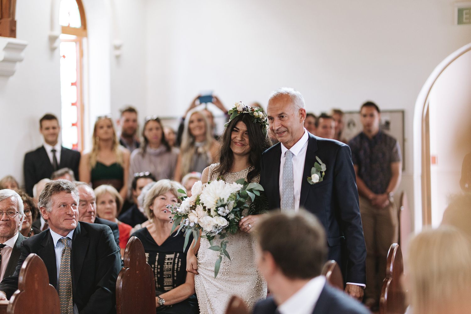 Wedding guests seated in church pews watch ceremony with emotional expressions during intimate indoor celebration.