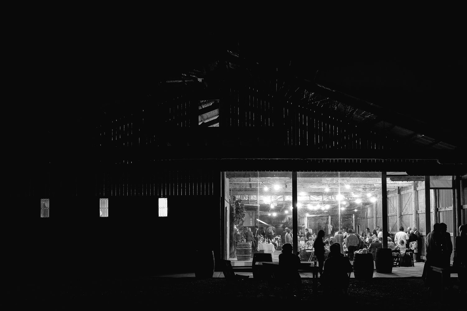 A black and white nighttime photograph shows silhouettes of people gathering inside an illuminated rural storefront.