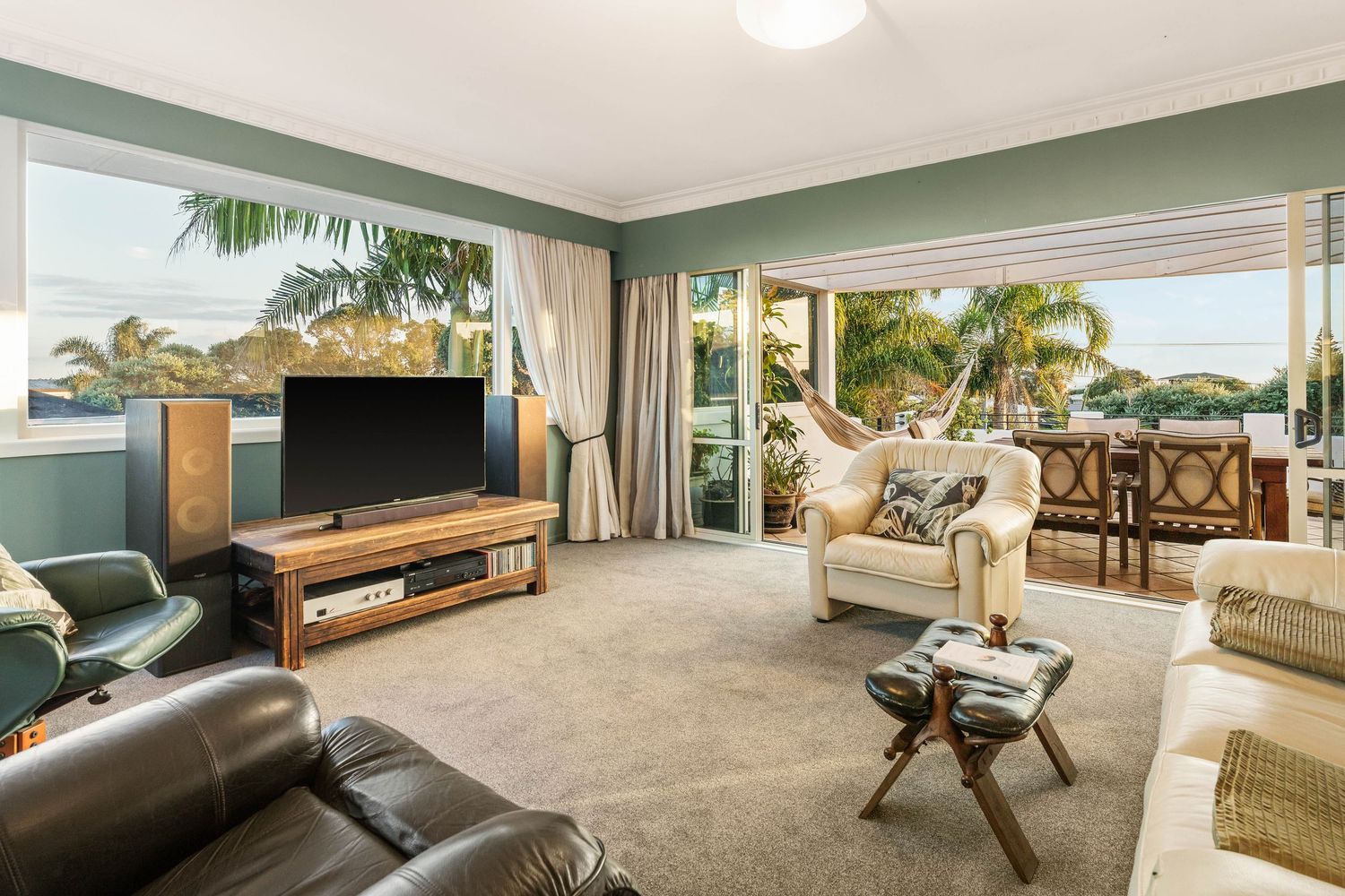 Spacious coastal living room with leather couches and TV setup overlooking palm trees through large windows.