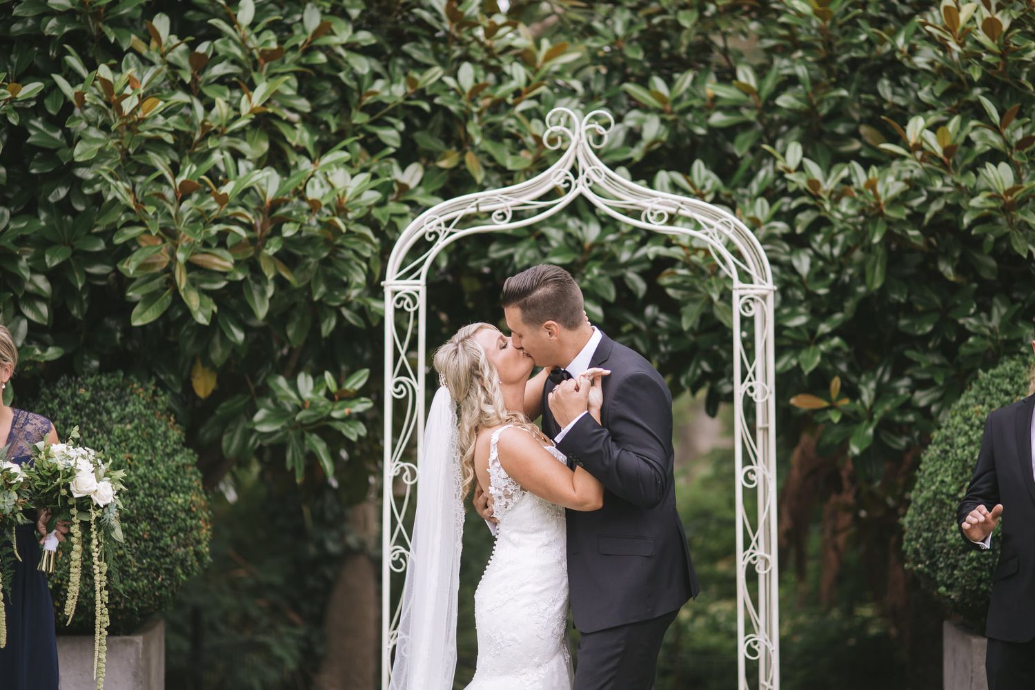 A romantic moment under an ornate white metal garden arch decorated with wedding florals against lush greenery.