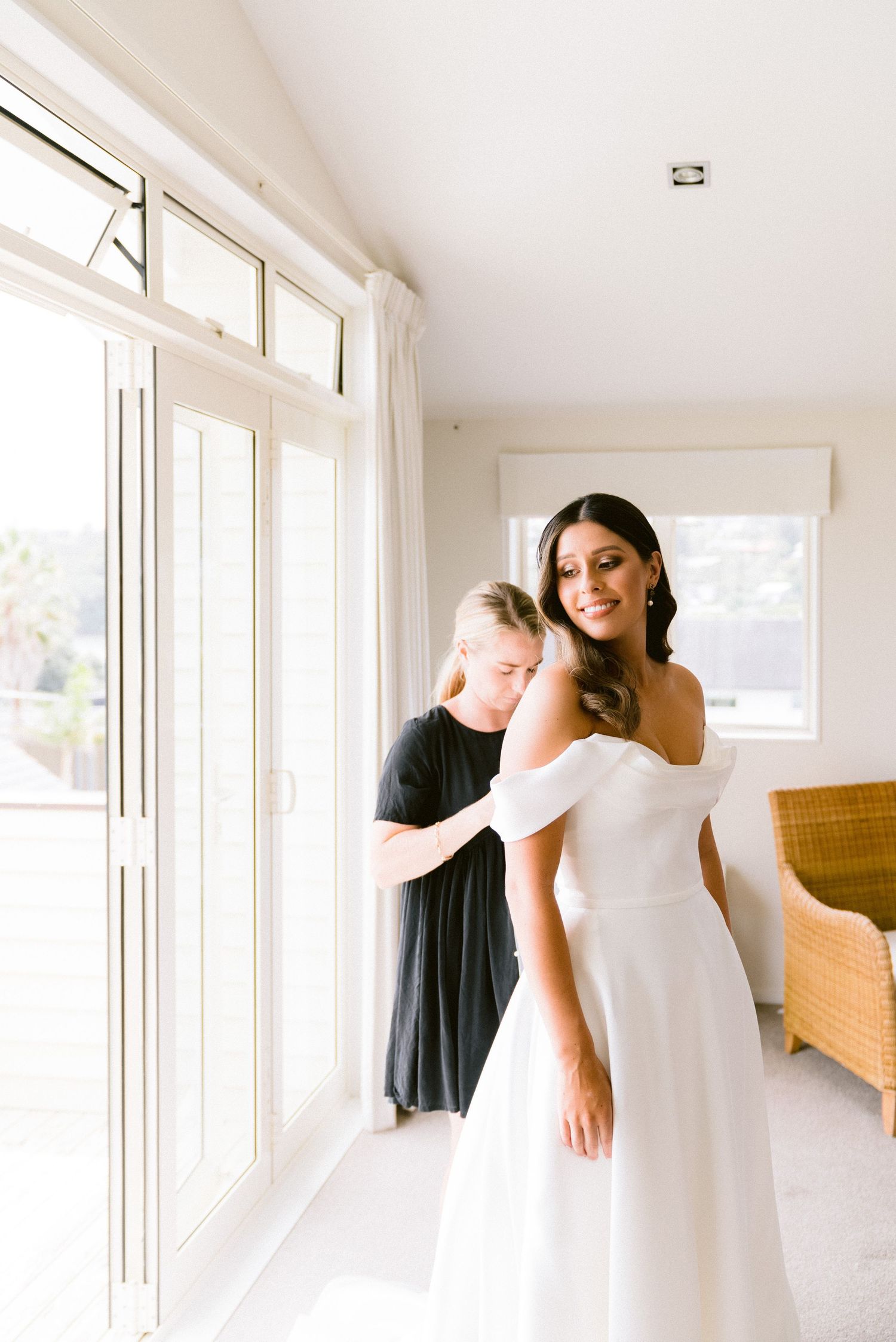 A wedding dress fitting moment in a bright sunlit room with an elegant off-shoulder white gown.