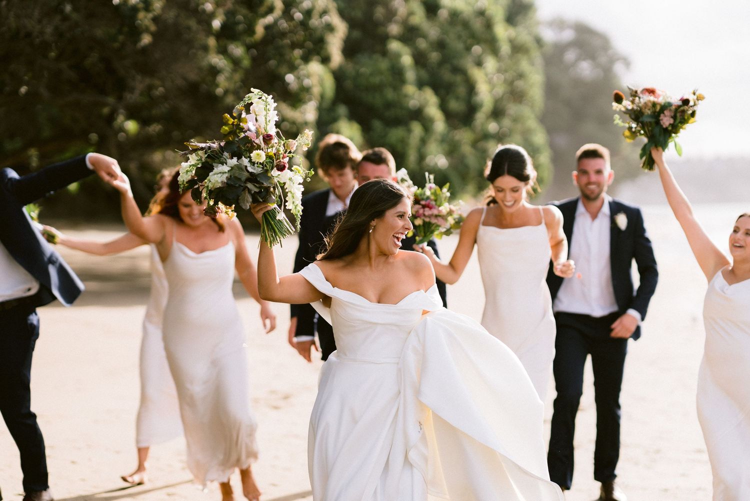 A wedding party joyfully walks together on a path with bridesmaids in white dresses and groomsmen in black suits.