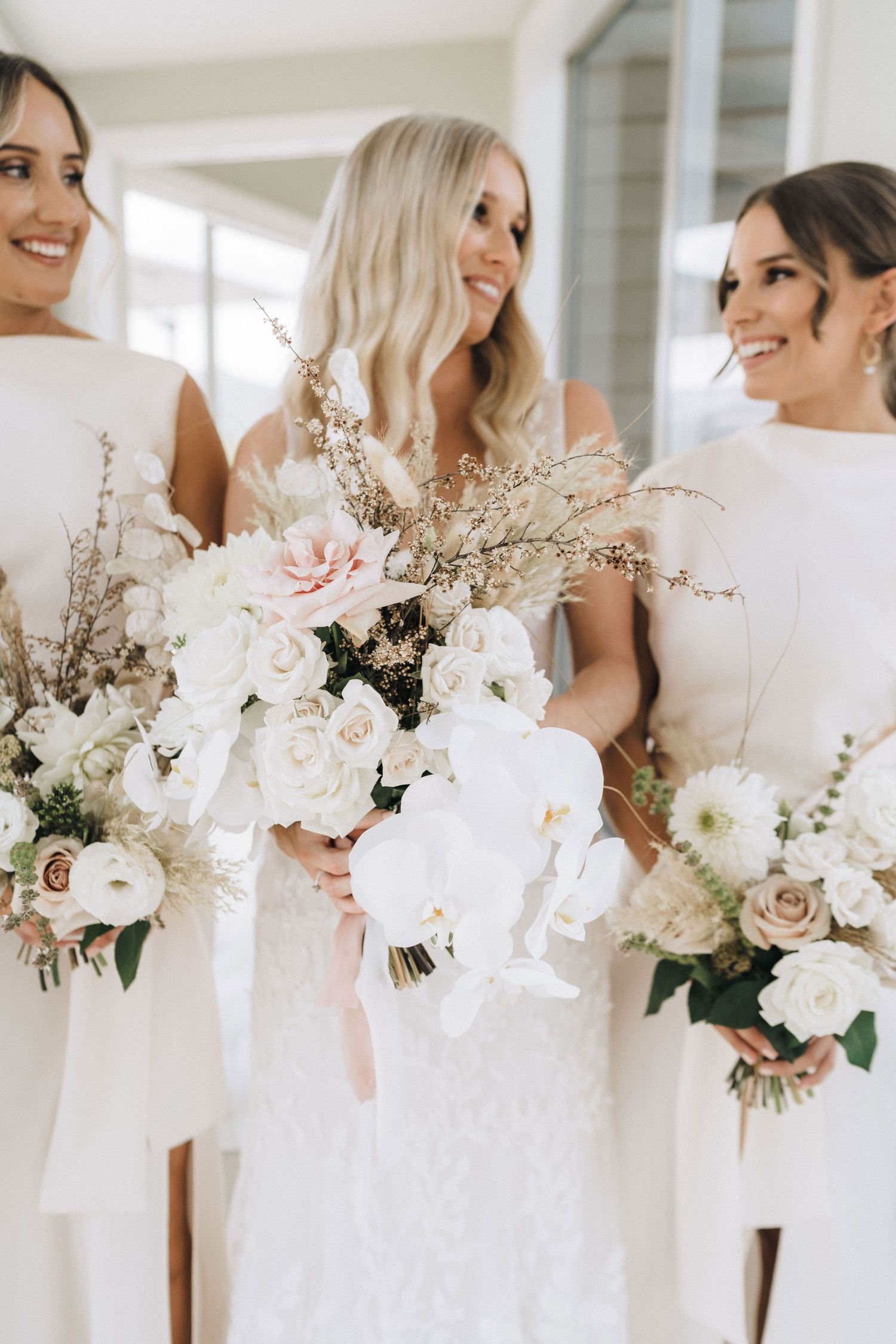 Three bridesmaids wearing white dresses hold elegant bouquets of blush pink and white flowers with delicate branches.