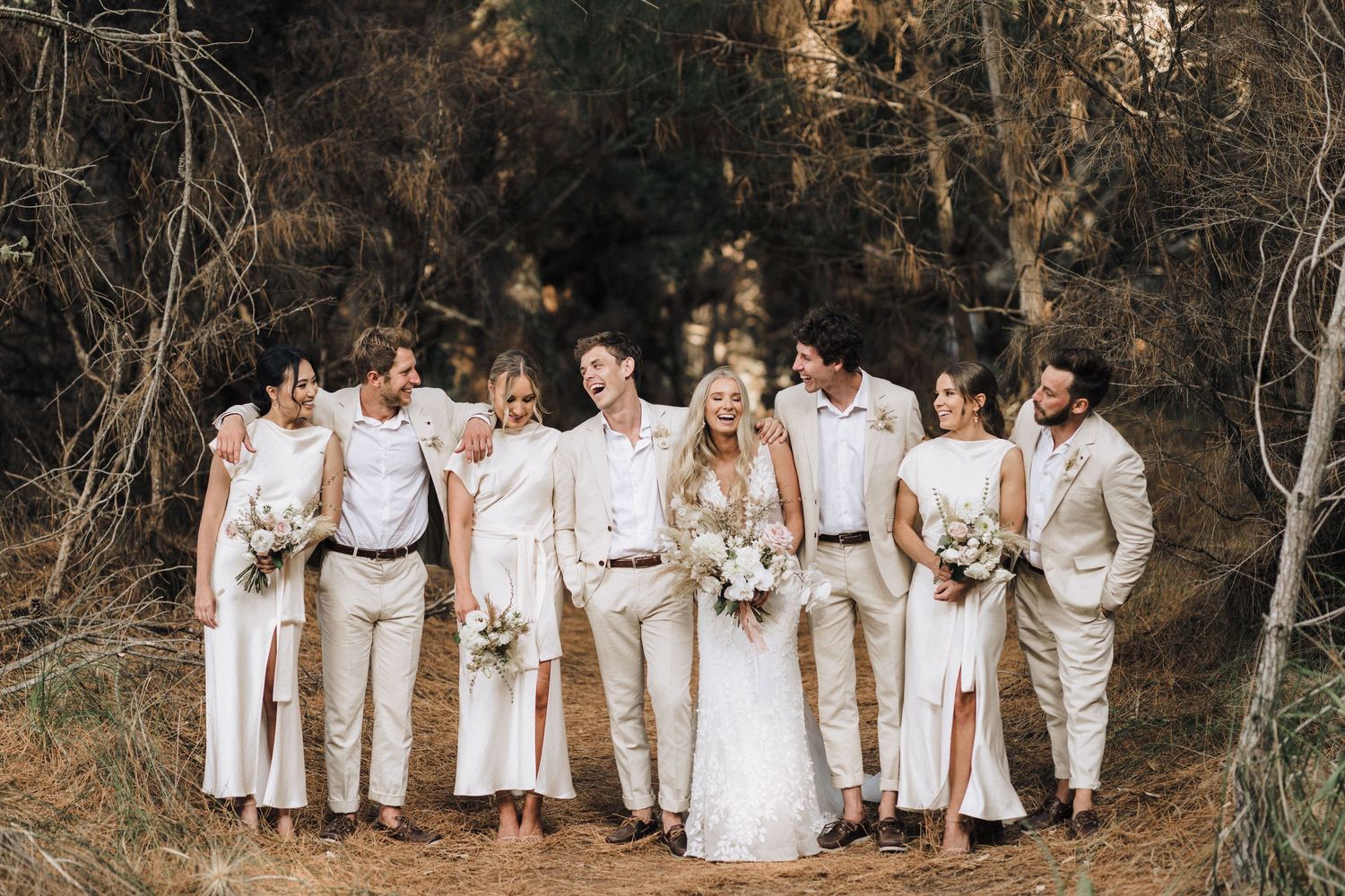 Wedding party in neutral beige and white outfits poses together in rustic outdoor forest setting.