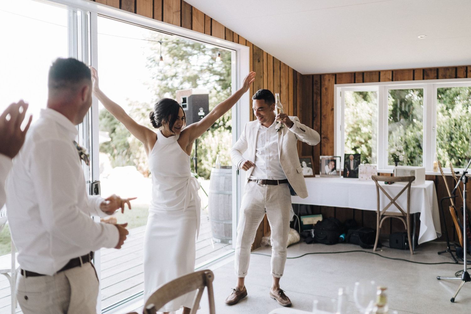 Wedding celebration with people dancing in white attire inside wooden-paneled reception room.