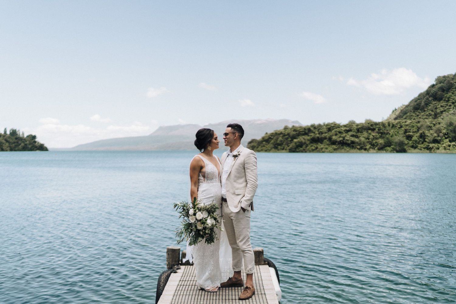 Romantic waterfront wedding portrait with couple embracing against serene lake and mountain backdrop.