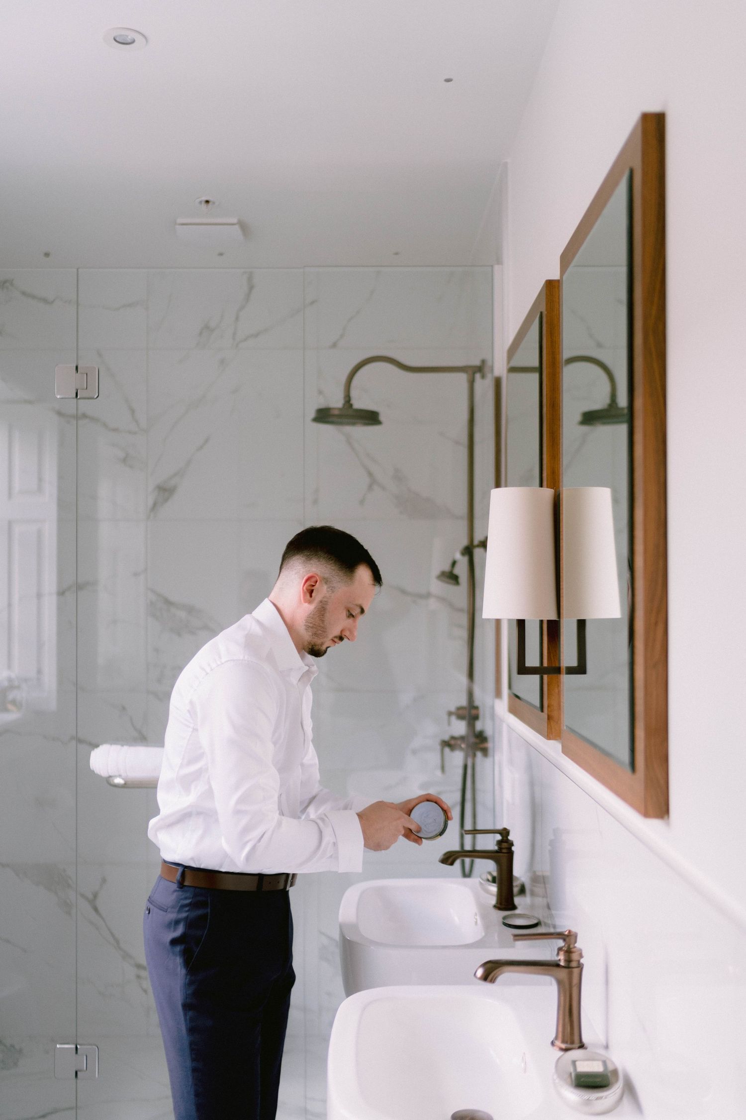 Person in white dress shirt and dark pants washing hands at modern white marble bathroom with brass fixtures and mirrors.