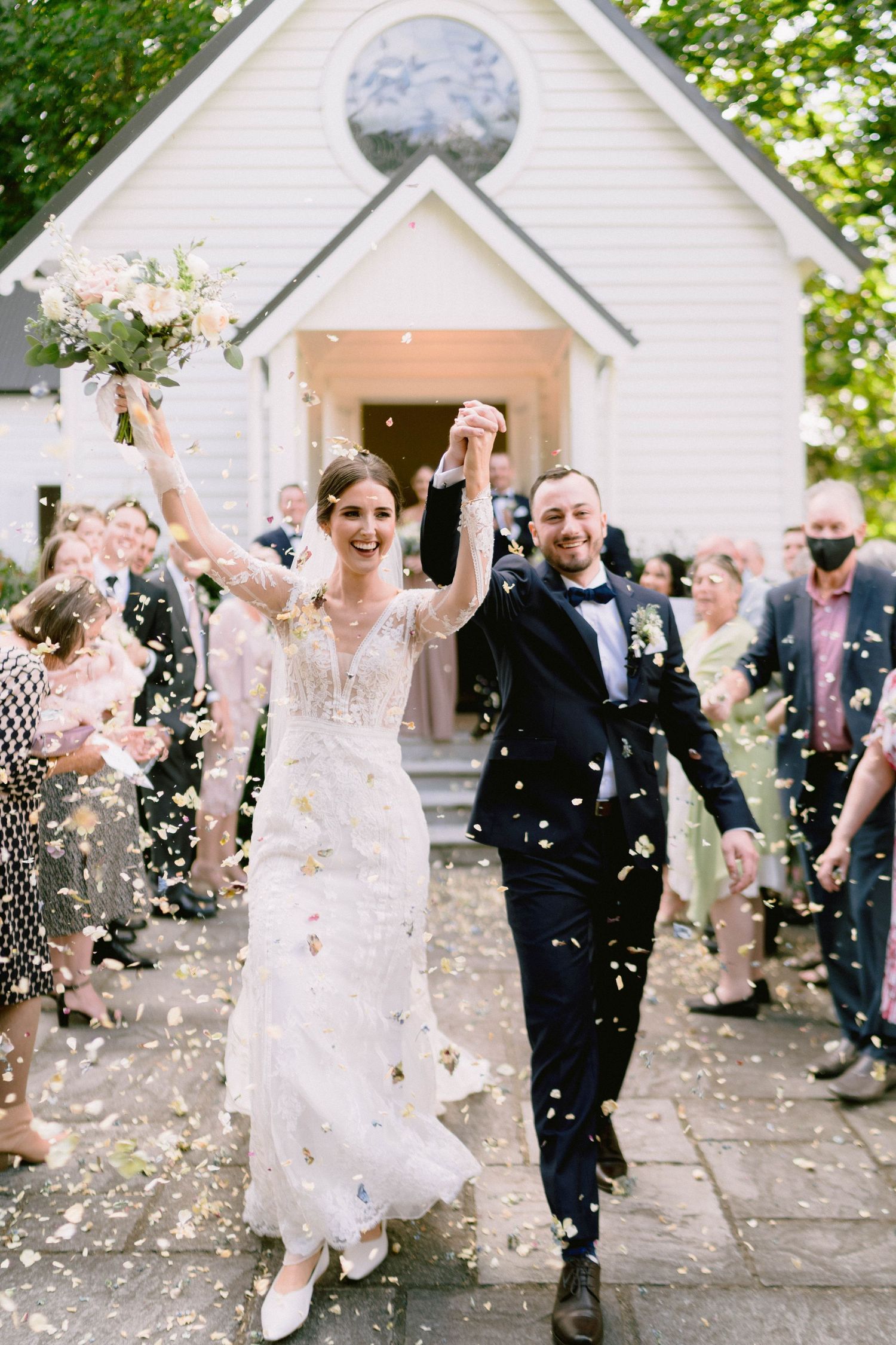 A just-married couple exits their wedding chapel while guests shower them with confetti and petals.