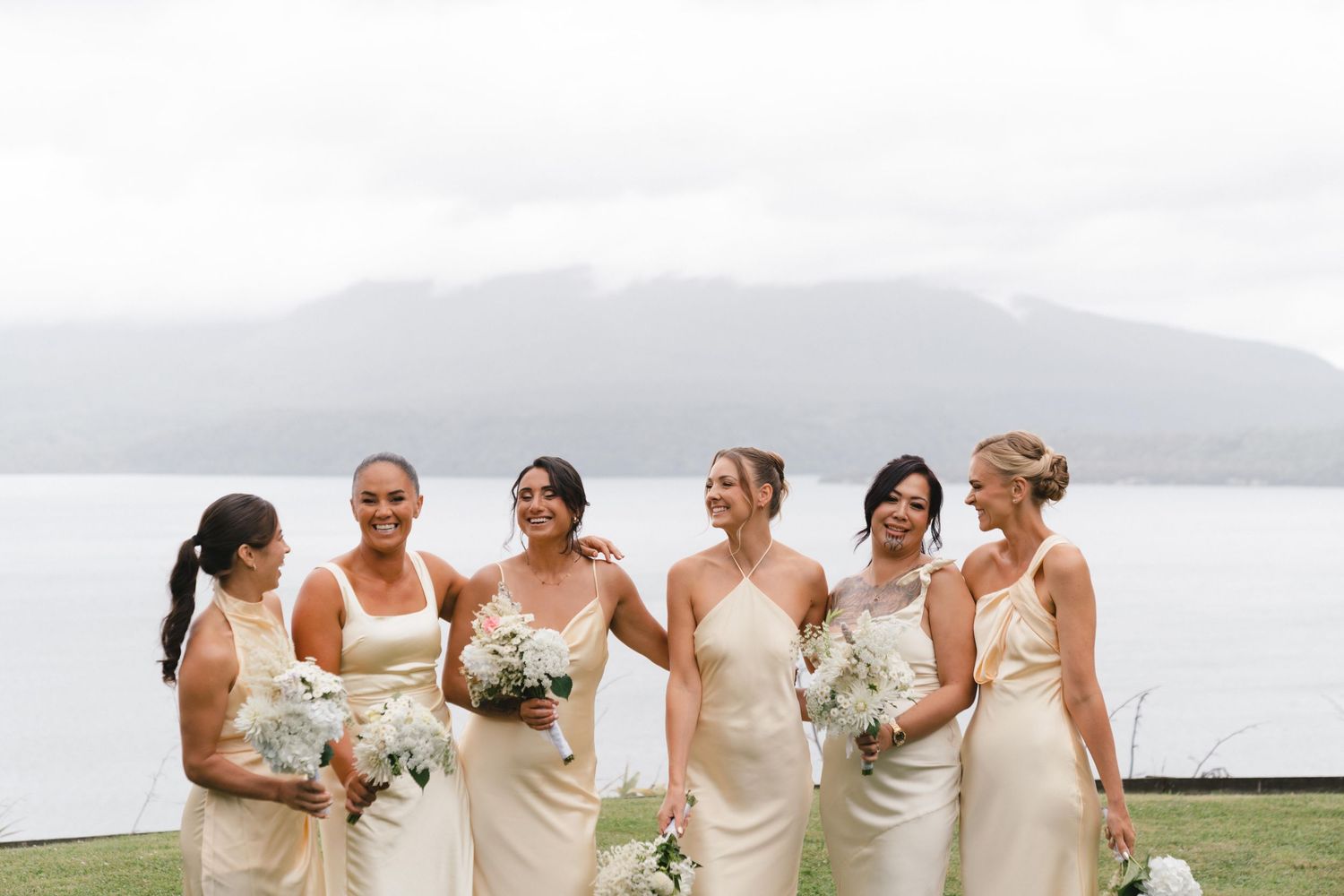 A group of bridesmaids in pale yellow dresses hold white bouquets while standing by a misty waterfront.
