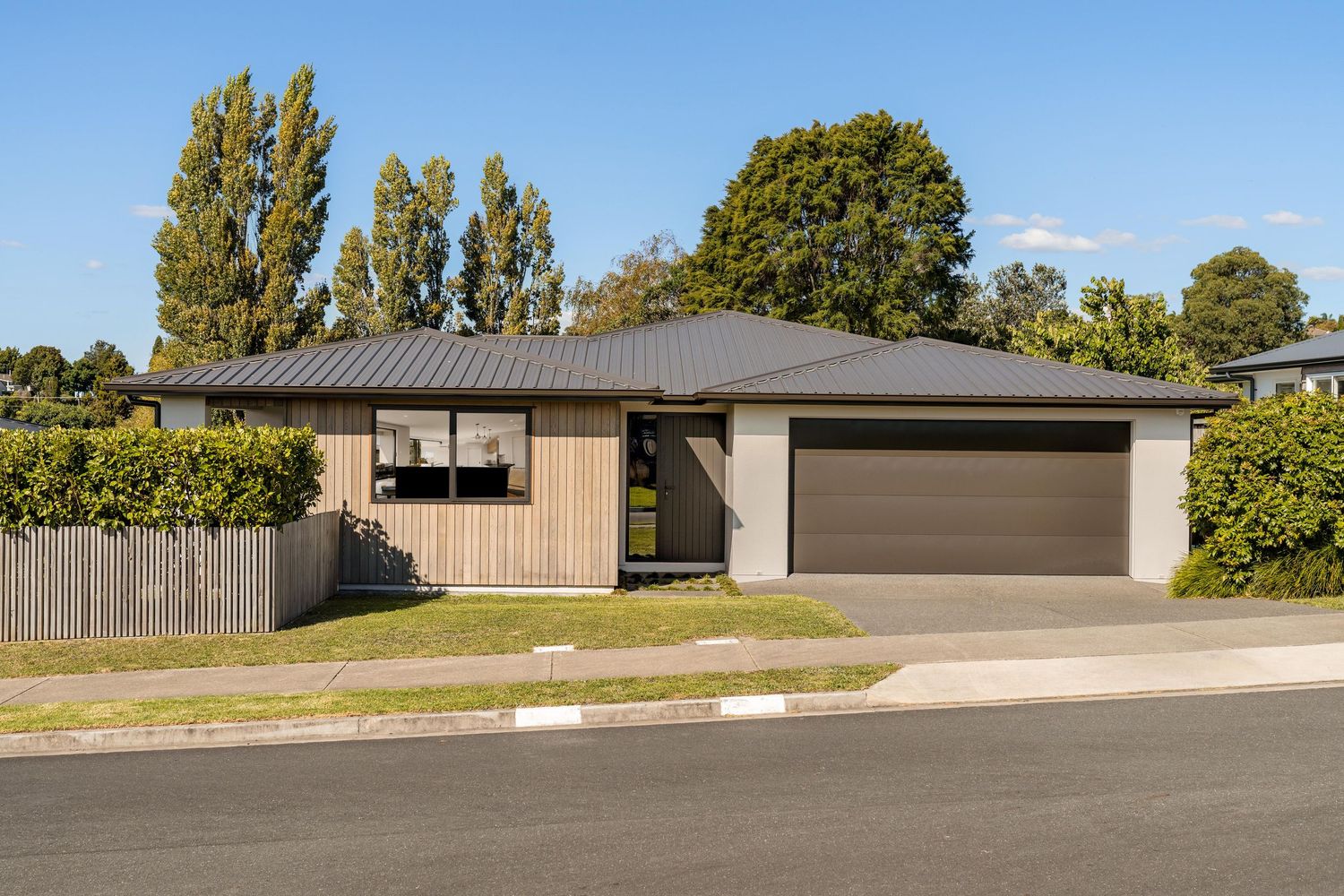 Modern single-story house with brown exterior, double garage and tall poplar trees in the background.
