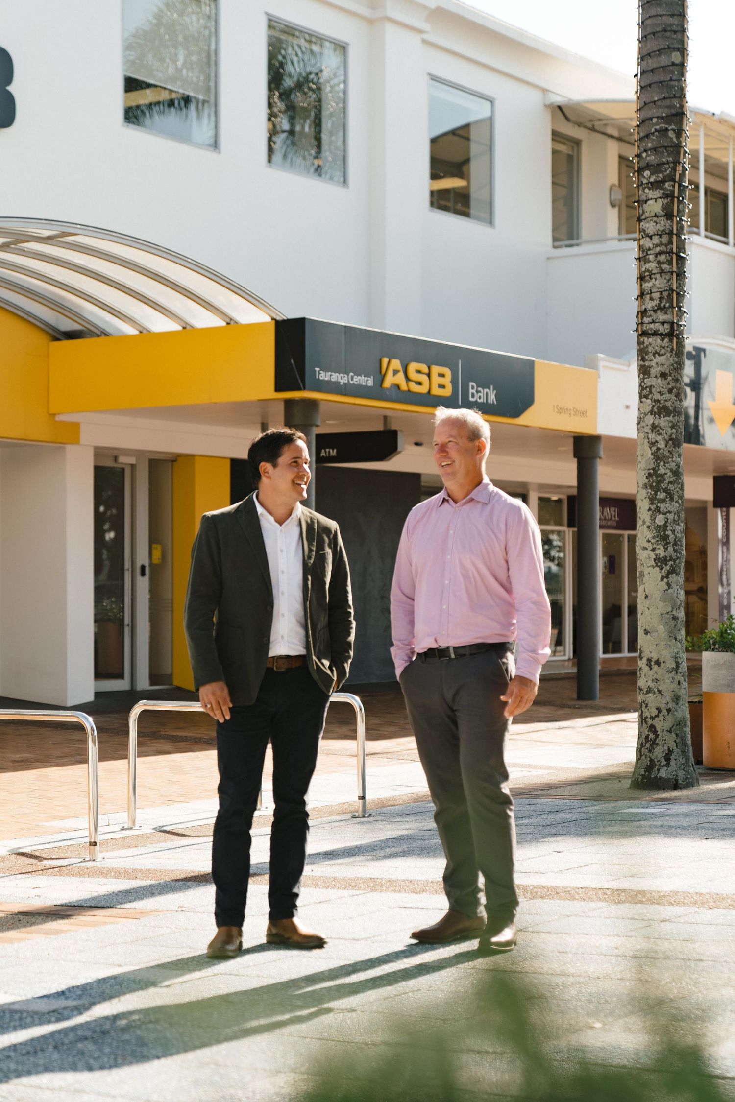 Two colleagues having conversation outside ASB Bank building entrance on sunny day.