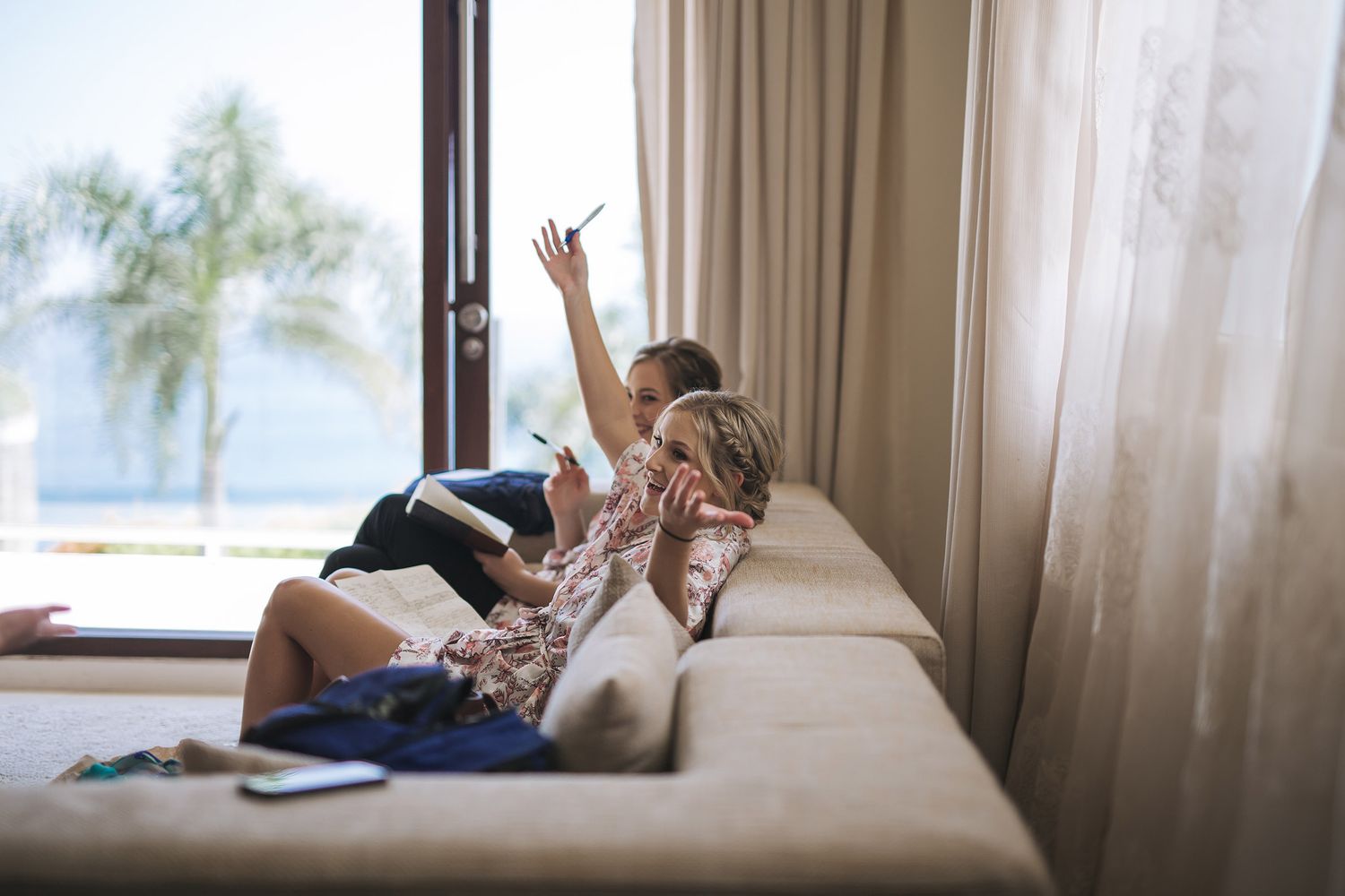 Young adult lounges playfully on beige couch by window with beach view in background.